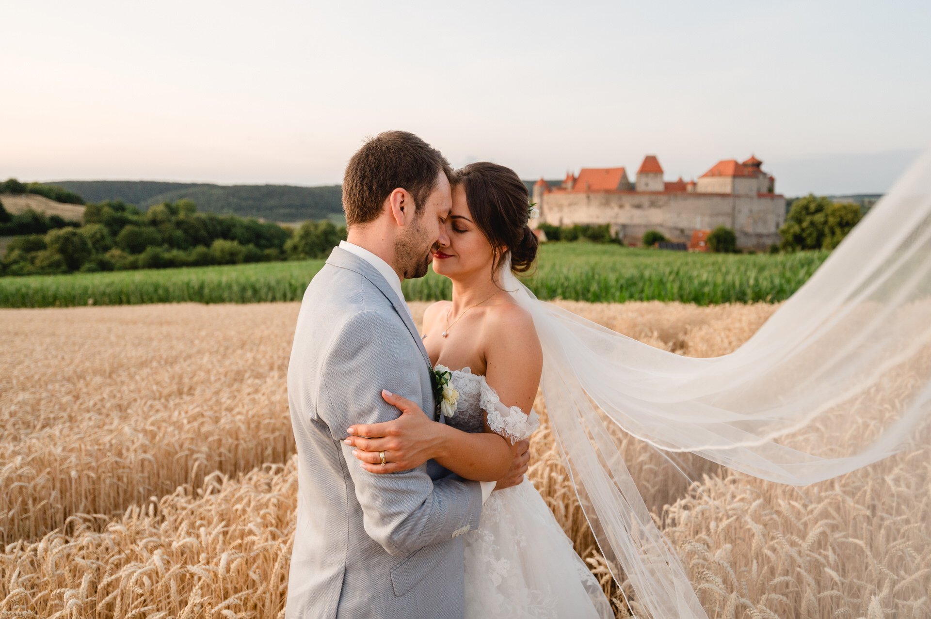 Braut und Bräutigam umarmen sich in einem Weizenfeld, im Hintergrund ist ein Schloss zu sehen. Der Schleier der Braut weht im Wind.