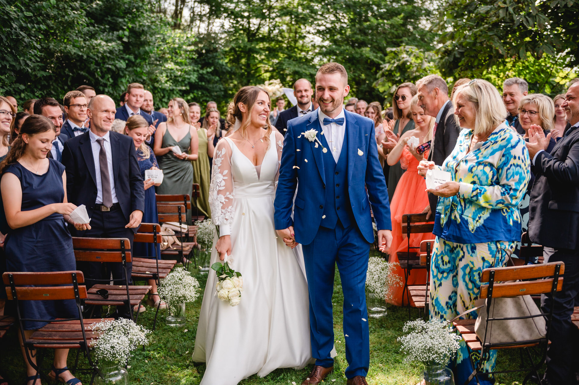 Ein Brautpaar lächelt, während es im Freien, umgeben von applaudierenden Gästen, den Gang entlanggeht.
