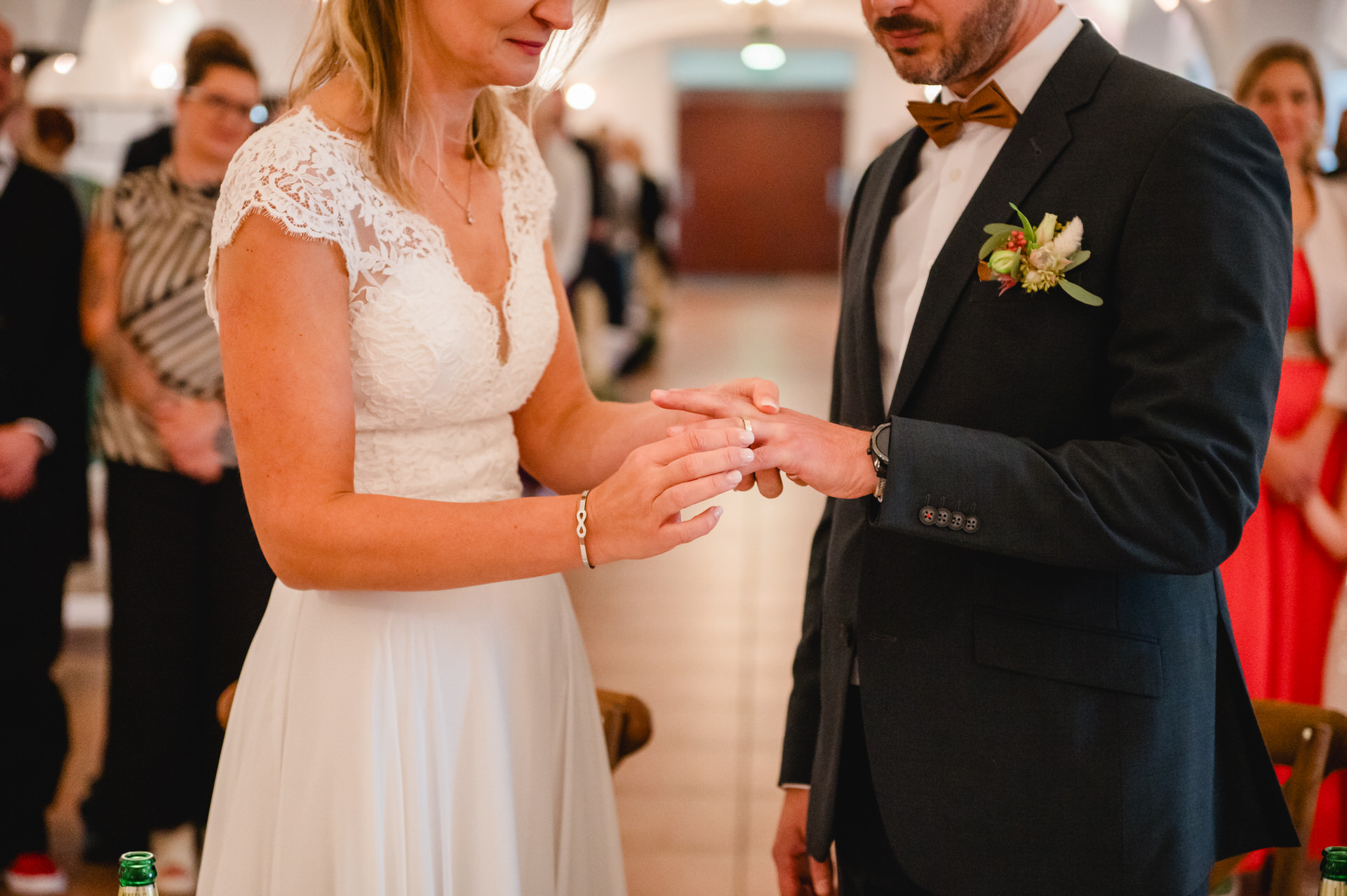 Während einer Hochzeitszeremonie steckt eine Braut dem Bräutigam einen Ring an den Finger. Sie stehen im Haus, umgeben von Gästen.