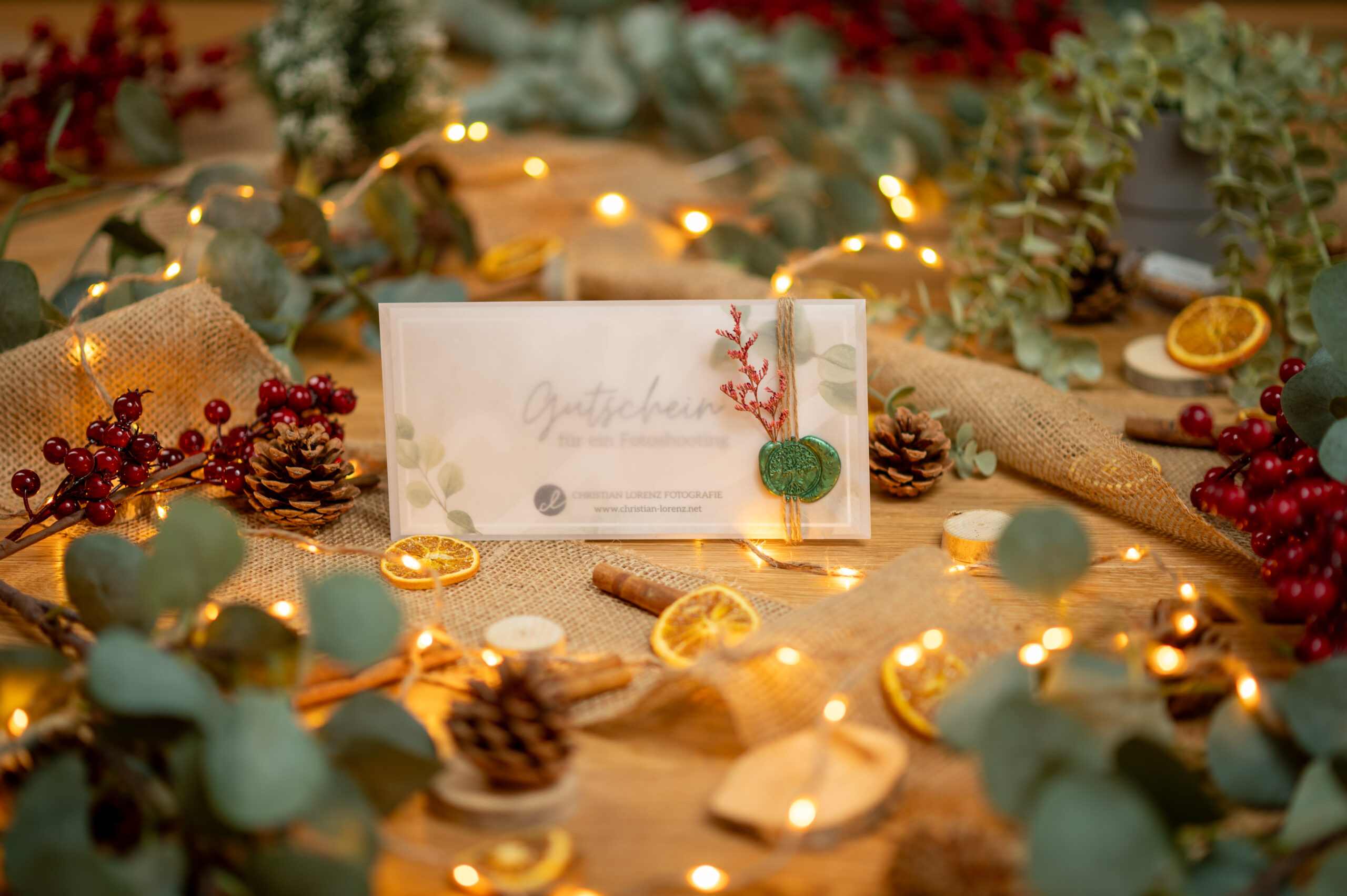 Eine festliche Geschenkkarte, umgeben von Tannenzapfen, Orangenscheiben, Zimtstangen, Beeren, Eukalyptusblättern und Lichterketten auf einem mit Sackleinen bedeckten Tisch.