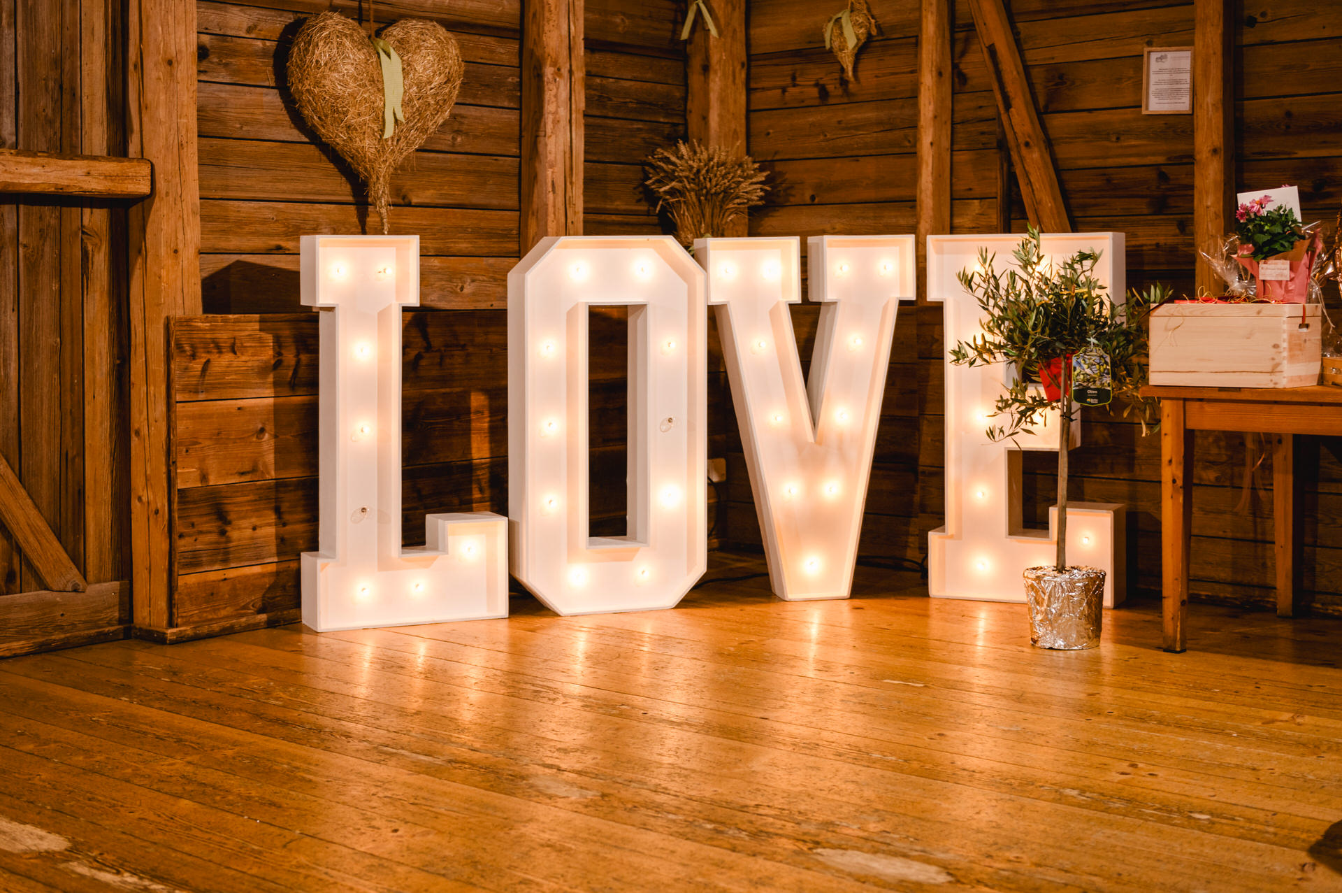Im charmanten, rustikalen Raum des Hochzeits-Schammenhofs in Langenau bei Ulm prangen leuchtende Festzeltbuchstaben mit der Aufschrift „LOVE“ stolz auf einem Holzboden, geschmückt mit einem Herzkranz und üppigem Grün.