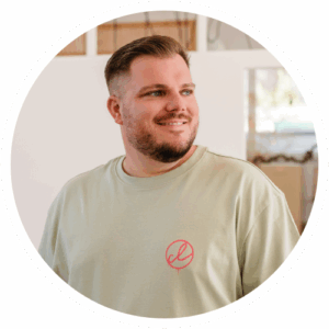 Ein Mann mit kurzen Haaren und Bart, der ein hellgrünes T-Shirt mit rotem Logo trägt, steht in einem Raum und schaut lächelnd nach links.