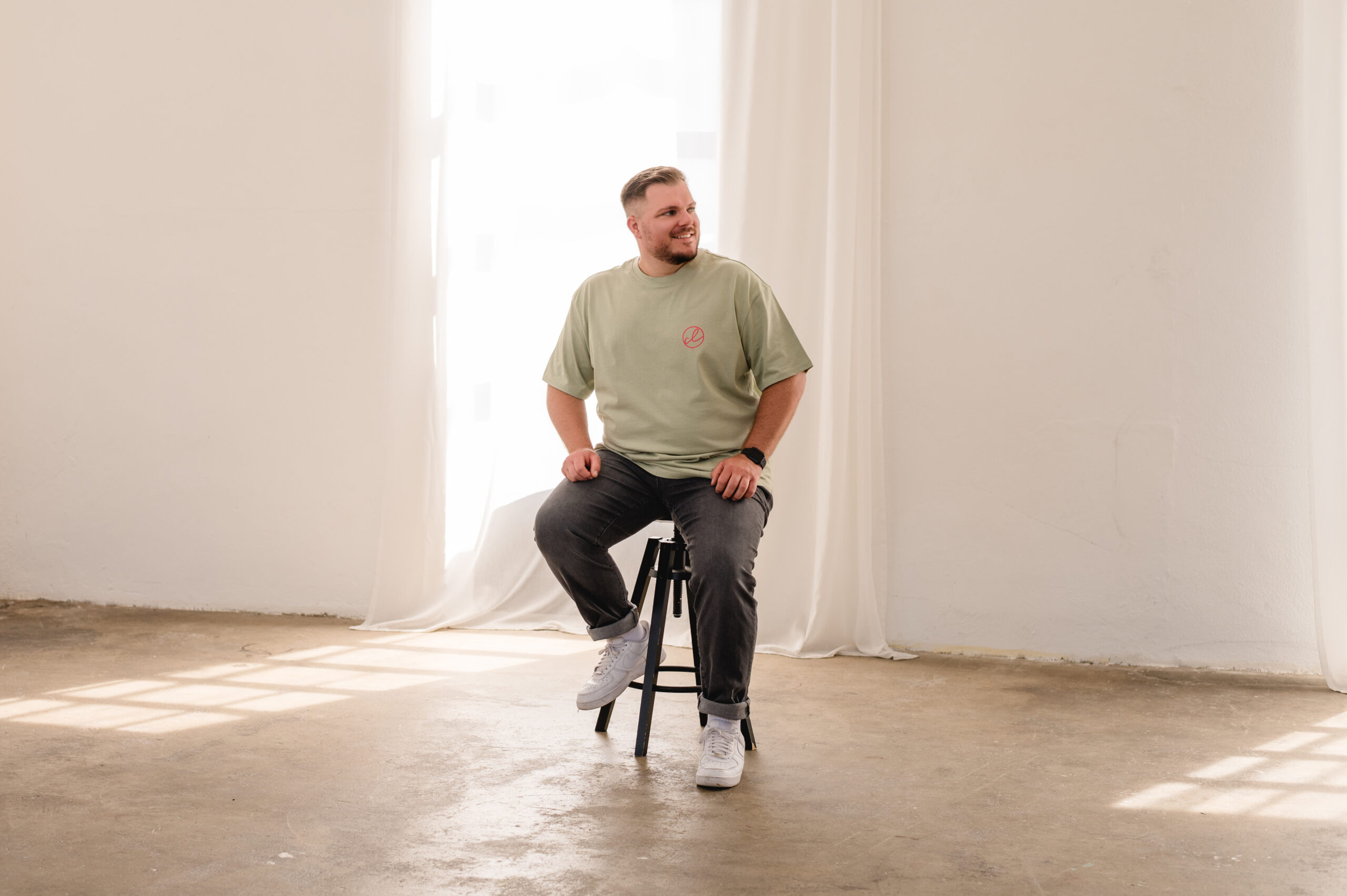 Ein Mann sitzt auf einem Hocker in einem hellen Raum mit weißen Vorhängen und einfallendem Sonnenlicht und denkt in aller Ruhe über den Auszug Trauung nach.