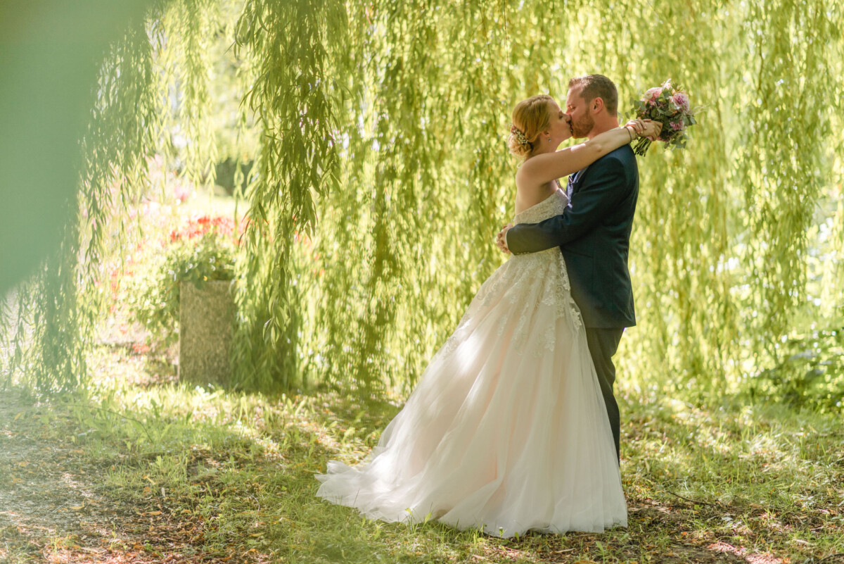 Natalie_Willi_Sulzemoos_00030 ein Brautpaar, das sich unter einer Weide küsst.