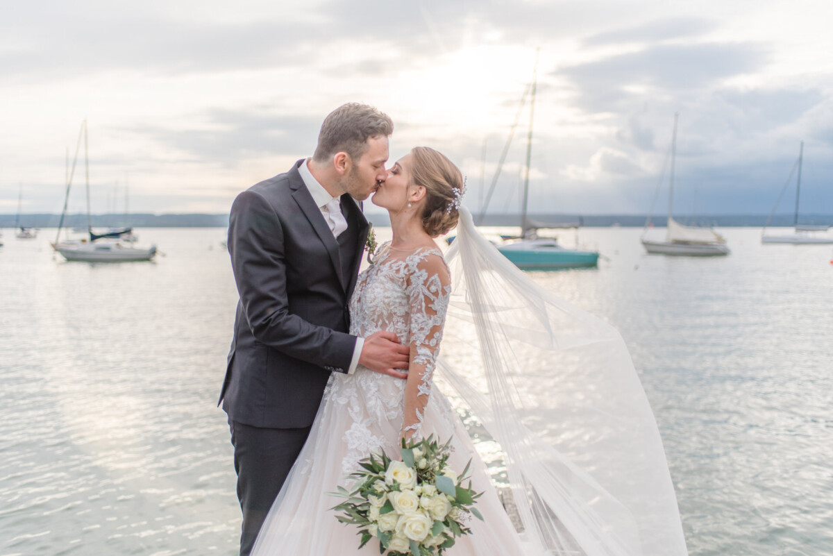 Sophia_Riccardo_Ammersee_00023 eine Braut und ein Bräutigam, die auf einem Dock mit Booten im Hintergrund küssen.