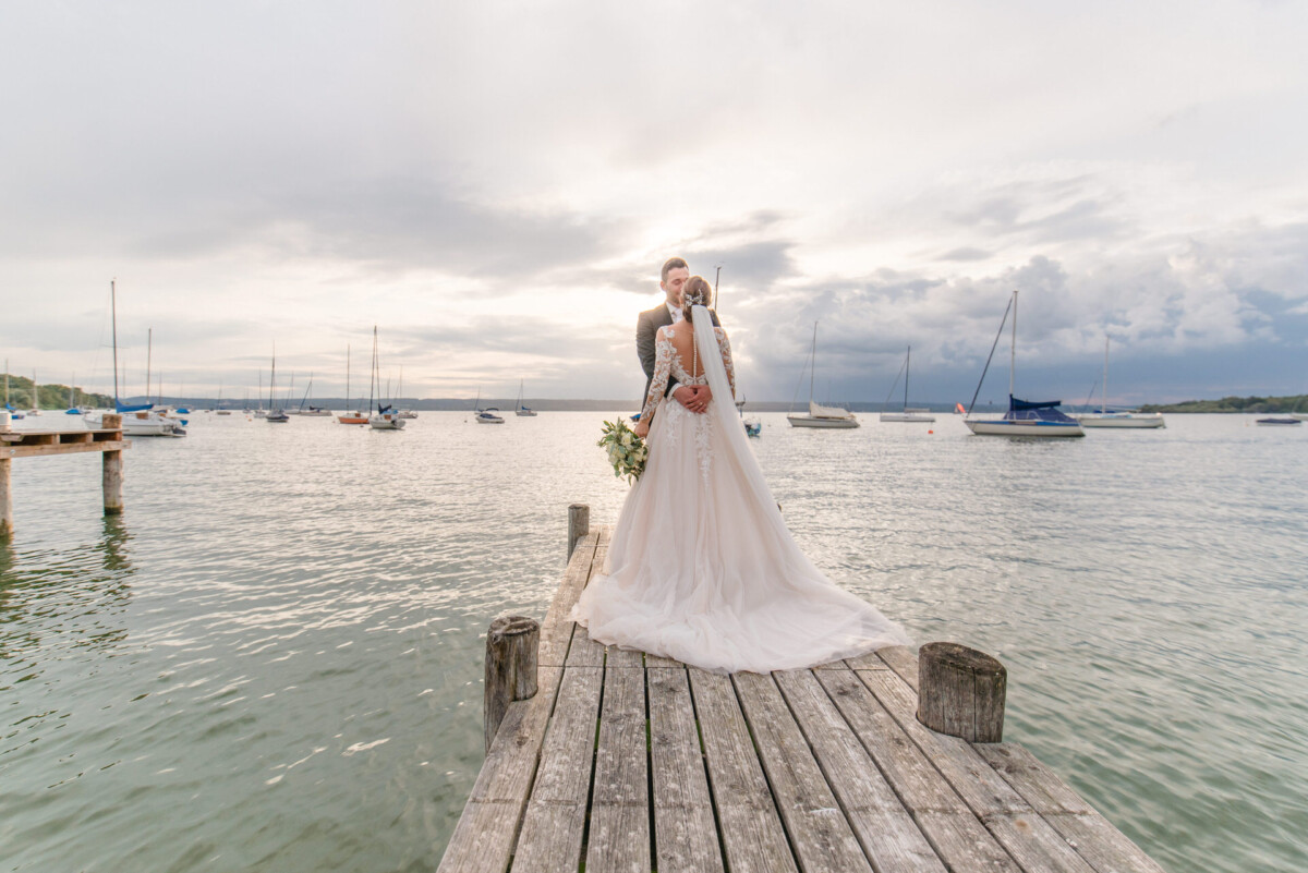 Sophia_Riccardo_Ammersee_00027 Eine Braut und ein Bräutigam stehen auf einem Dock.