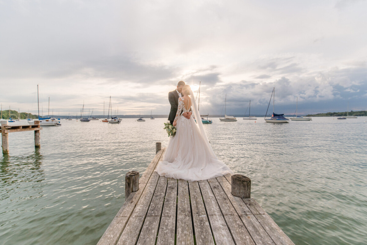 Sophia_Riccardo_Ammersee_00028 Eine Braut und ein Bräutigam stehen auf einem Dock.