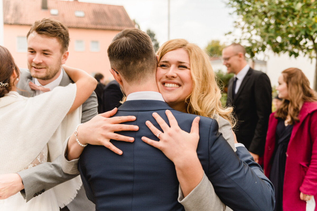 R_M_Bobingen_Langerringen_Hochzeit_00313_web Ein Mann und eine Frau umarmen sich vor einer Menschenmenge.