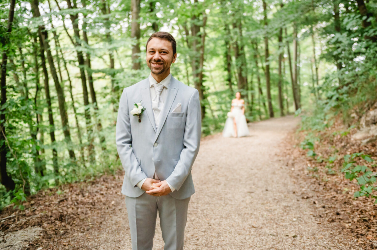Ein Mann im hellgrauen Anzug steht lächelnd auf einem Waldweg, von hinten nähert sich eine verschwommene Gestalt im Brautkleid.