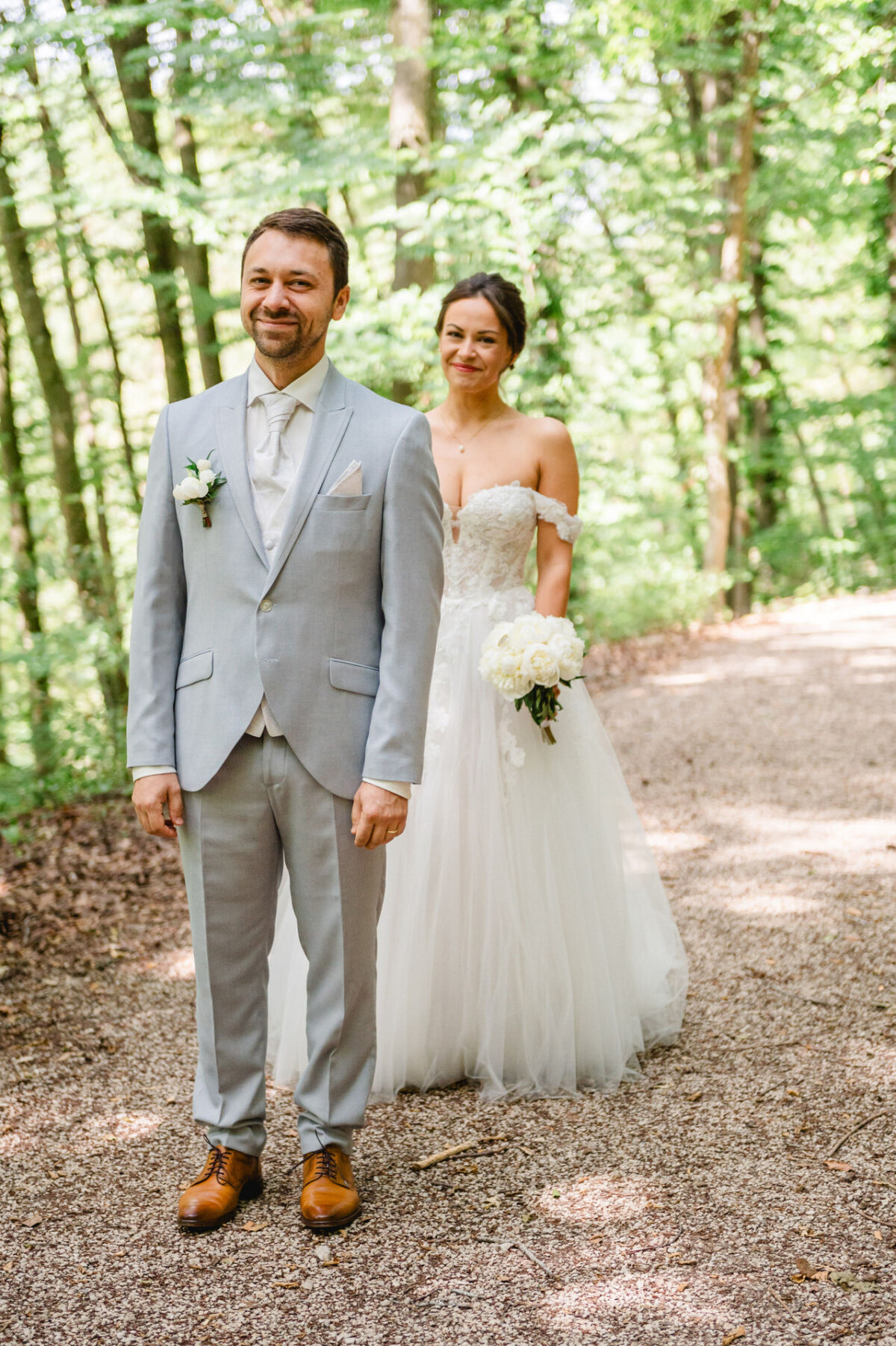 Ein Bräutigam im hellgrauen Anzug steht lächelnd da, während sich auf einem Waldweg eine Braut im weißen Kleid von hinten nähert.