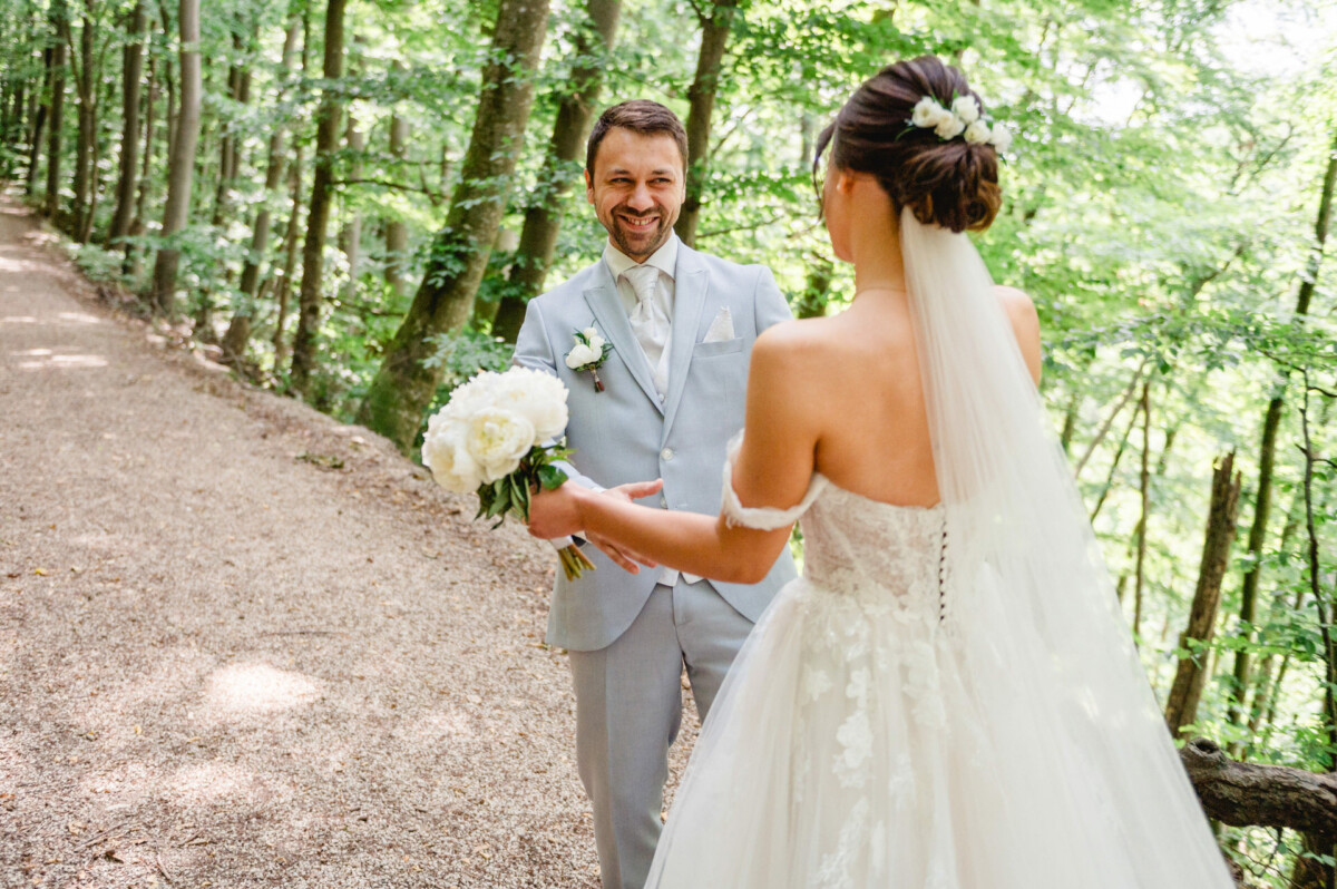 Braut und Bräutigam im Hochzeitskleid stehen auf einem Waldweg. Die Braut hält einen Strauß weißer Blumen in der Hand und blickt dem lächelnden Bräutigam entgegen.