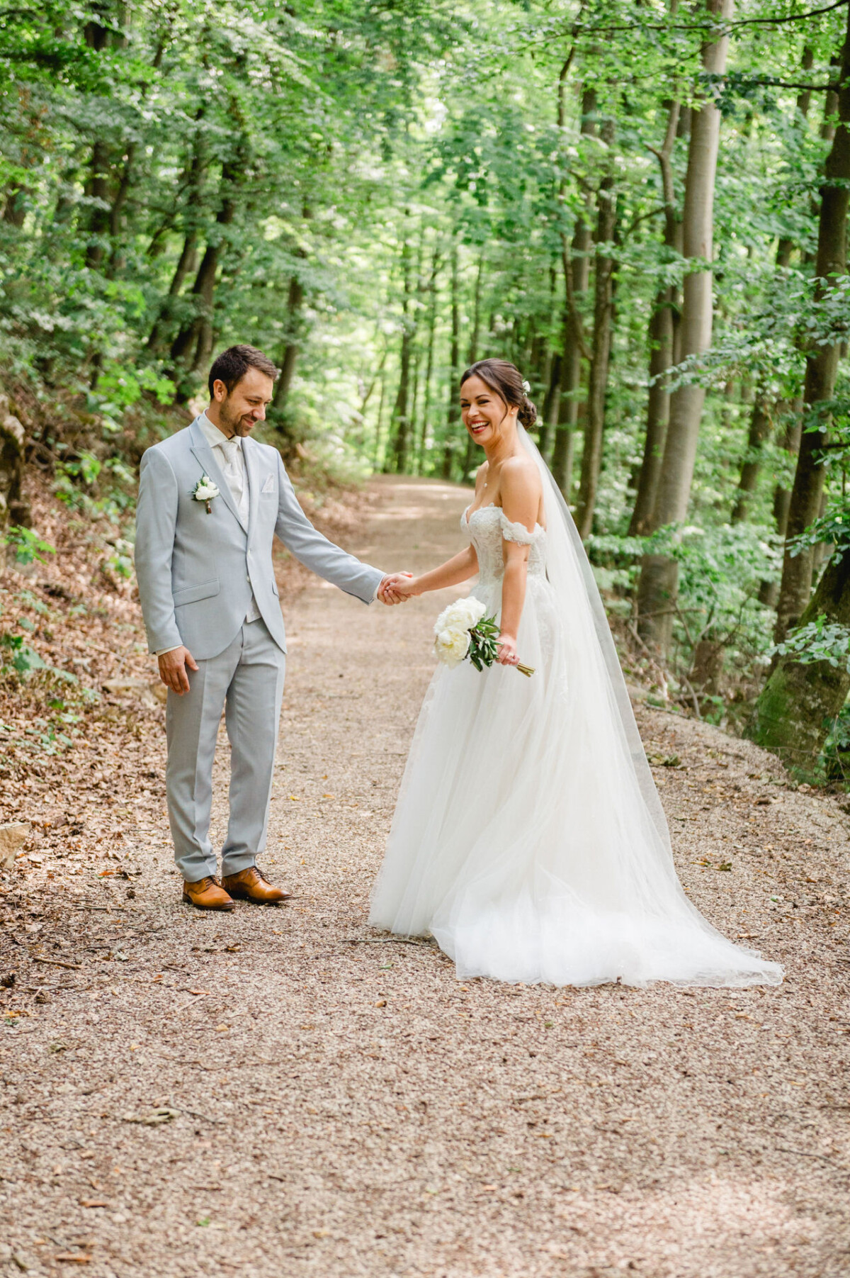Eine Braut im weißen Kleid und ein Bräutigam im hellgrauen Anzug halten auf einem Waldweg Händchen.