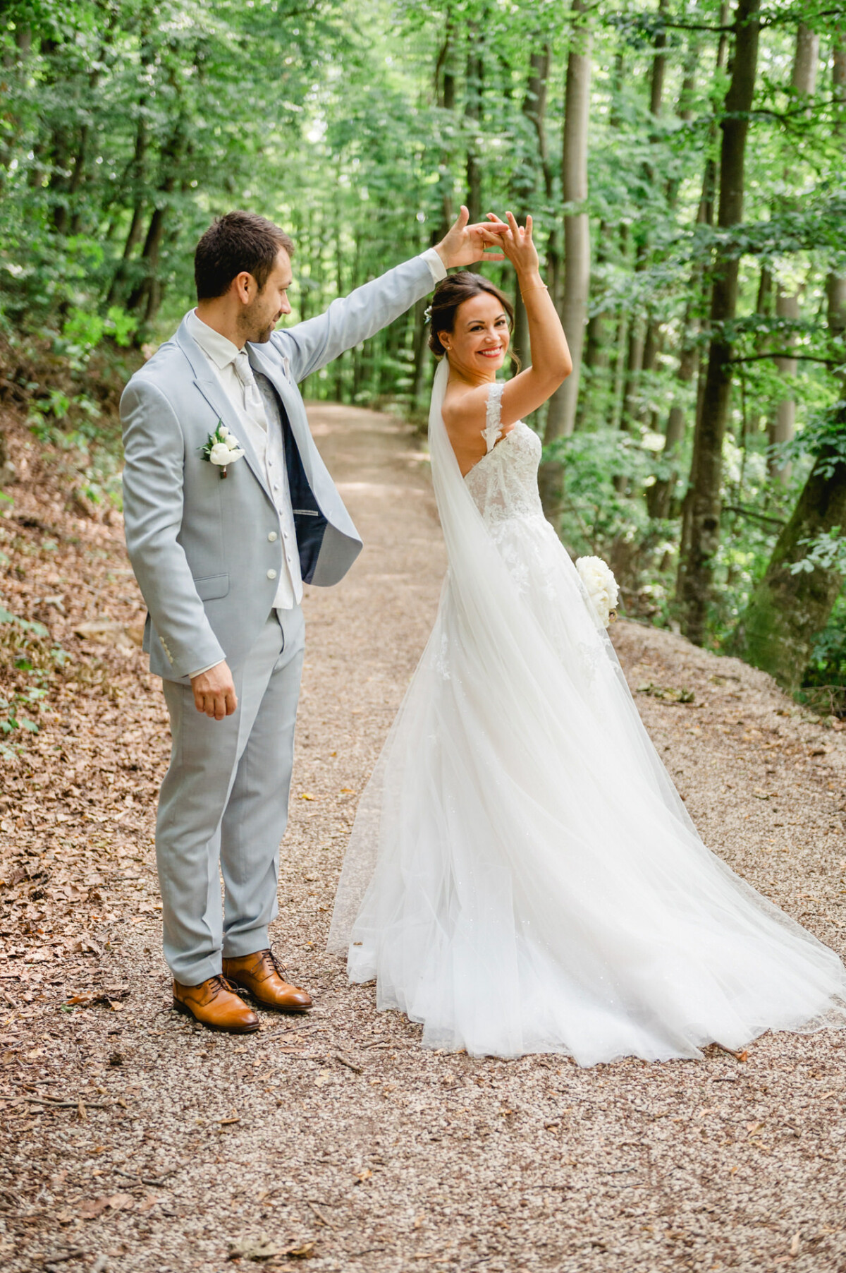 Braut und Bräutigam tanzen auf einem Waldweg. Sie trägt ein weißes Kleid und er einen hellgrauen Anzug.