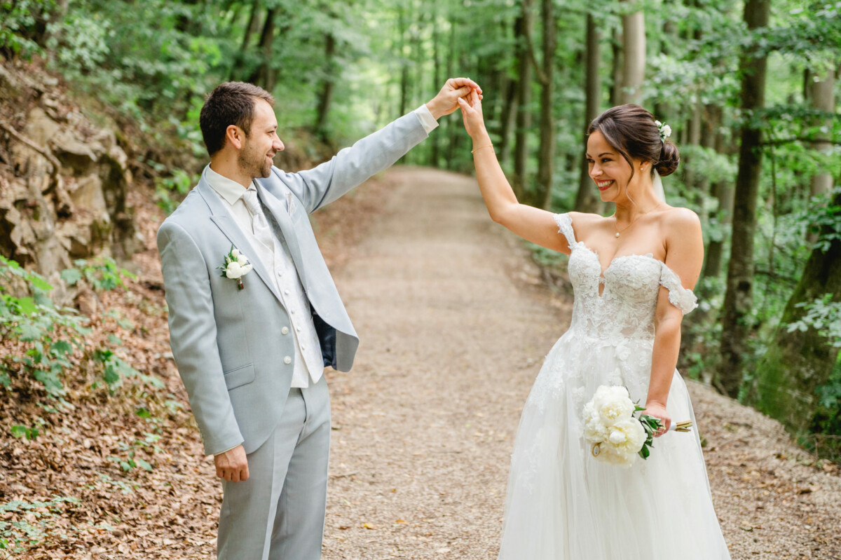 Ein Paar im Hochzeitskleid tanzt fröhlich auf einem Waldweg. Der Bräutigam trägt einen hellgrauen Anzug und die Braut ein weißes Kleid und hält einen Blumenstrauß.
