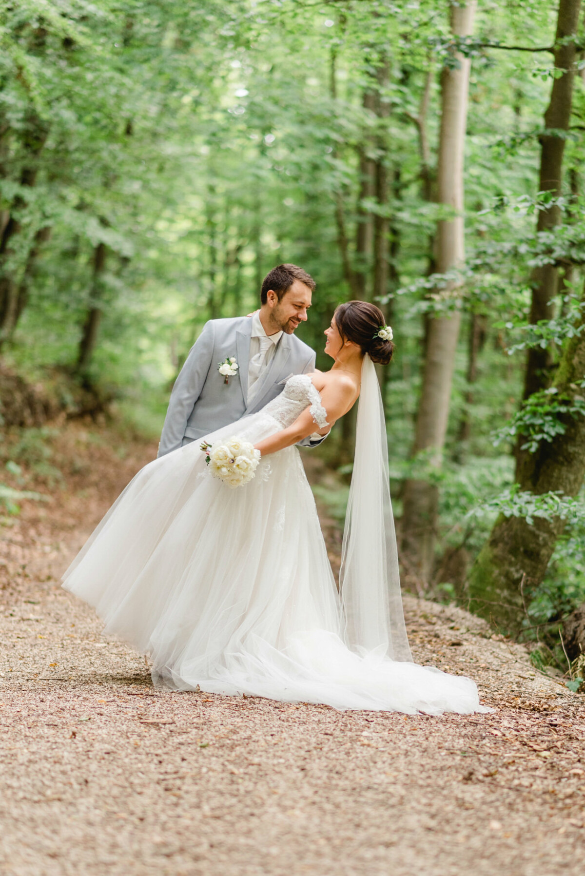 Ein Bräutigam im hellen Anzug und eine Braut im weißen Kleid verbringen einen freudigen Moment in einer üppigen, grünen Waldgegend.