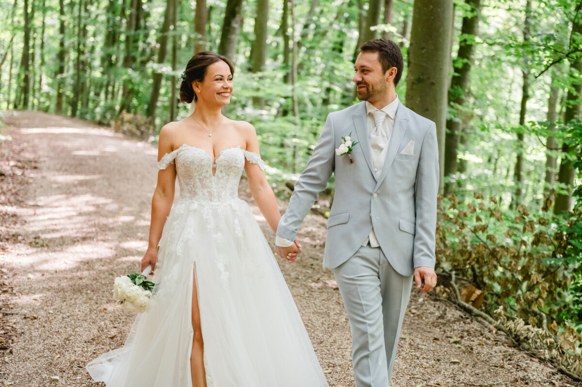 Braut und Bräutigam gehen Hand in Hand einen Waldweg entlang. Die Braut trägt ein weißes Kleid und hält einen Blumenstrauß in der Hand; der Bräutigam trägt einen hellgrauen Anzug. Beide lächeln, während sie sich anschauen.