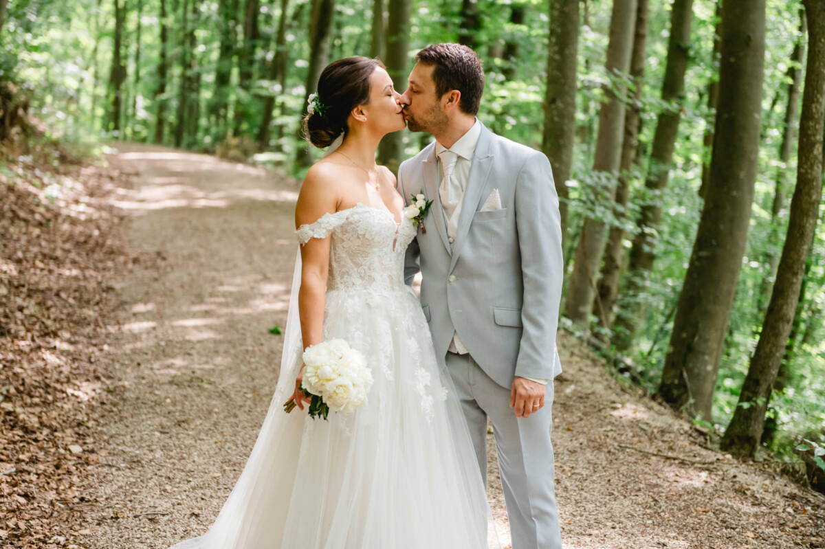 Ein Paar im Hochzeitskleid küsst sich auf einem Waldweg. Die Braut hält einen Strauß weißer Blumen.