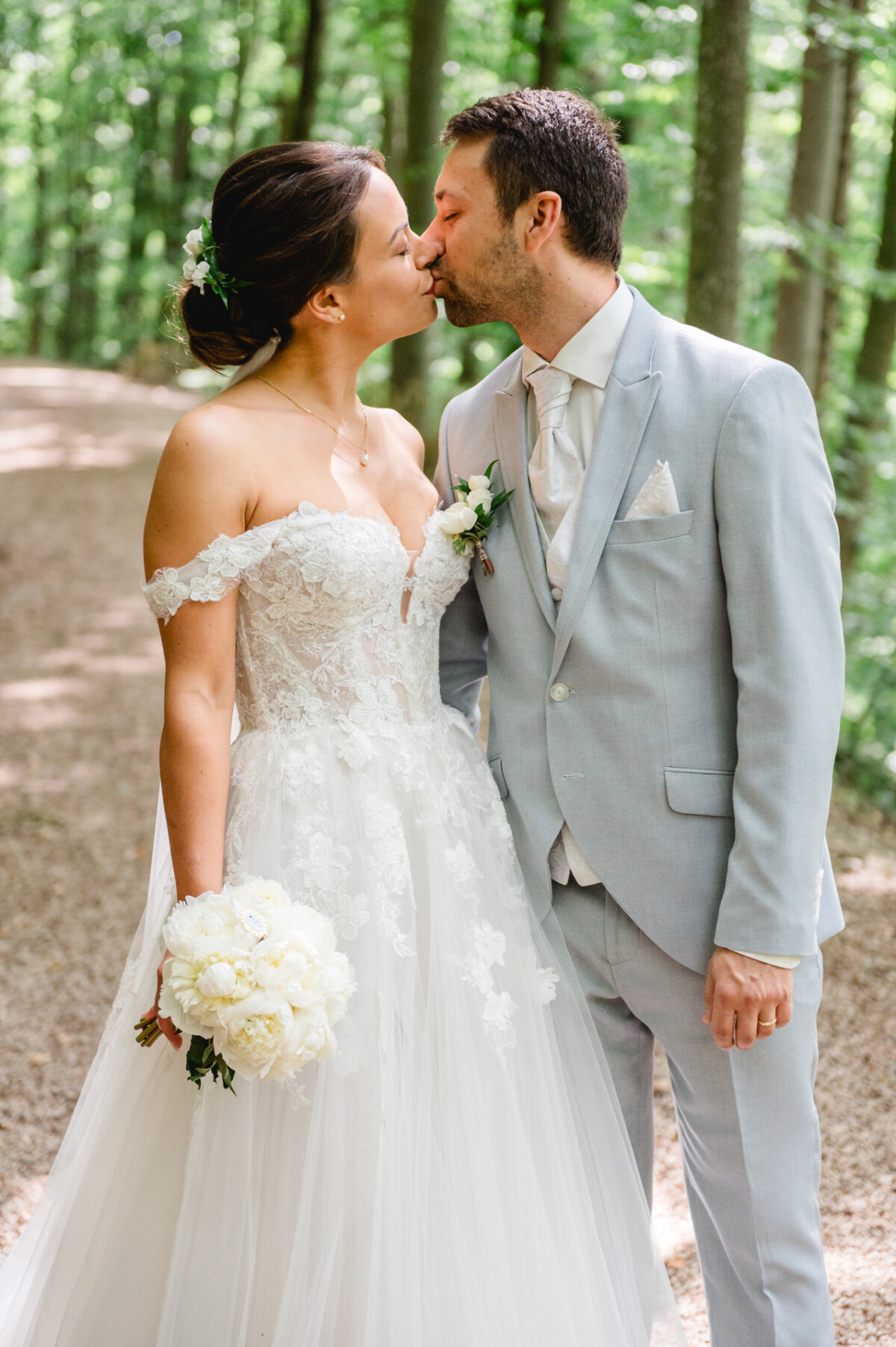 Ein Paar im Hochzeitskleid küsst sich auf einem Waldweg. Die Braut hält weiße Blumen in der Hand und trägt ein schulterfreies Spitzenkleid. Der Bräutigam trägt einen hellgrauen Anzug.