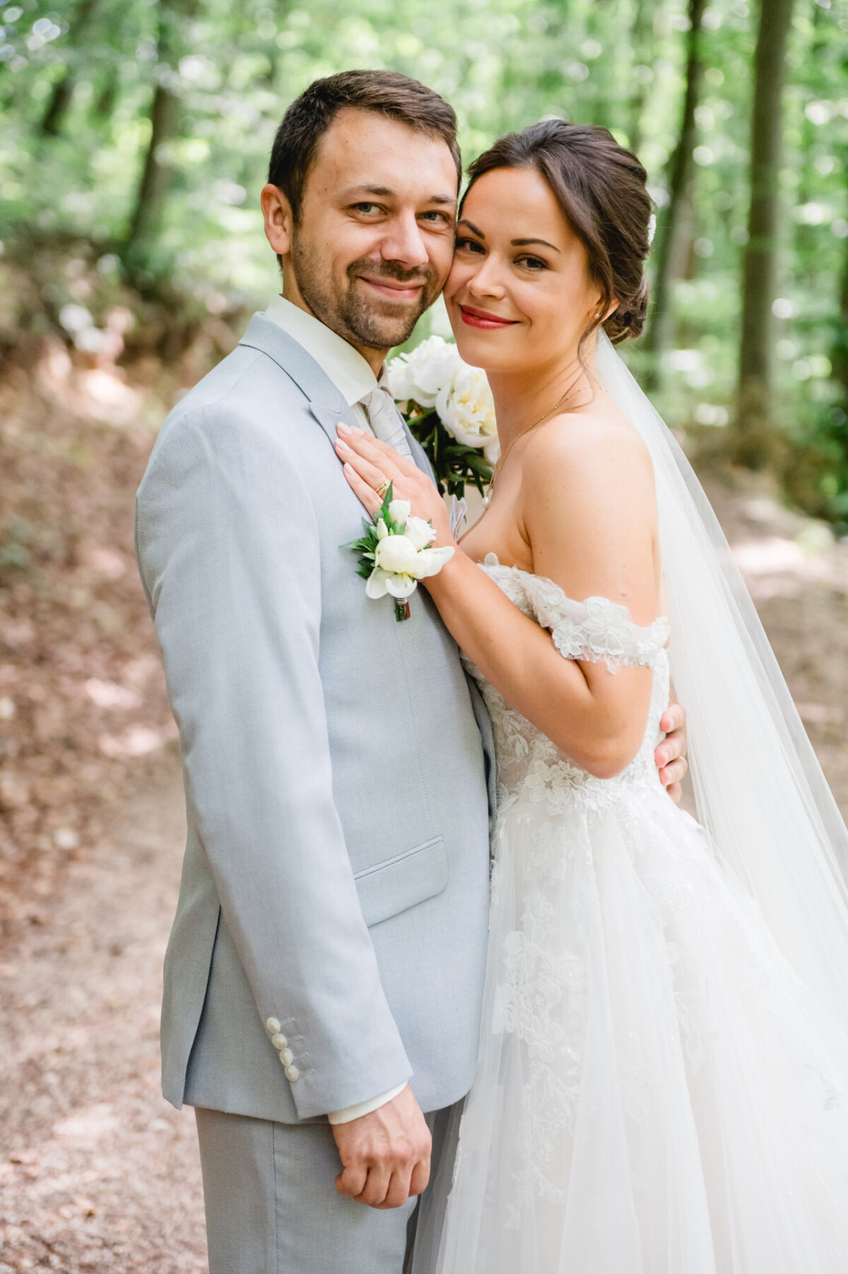 Braut und Bräutigam lächeln in einer Waldlandschaft. Die Braut trägt ein weißes Kleid und einen Schleier, der Bräutigam einen hellgrauen Anzug mit einer Ansteckblume.