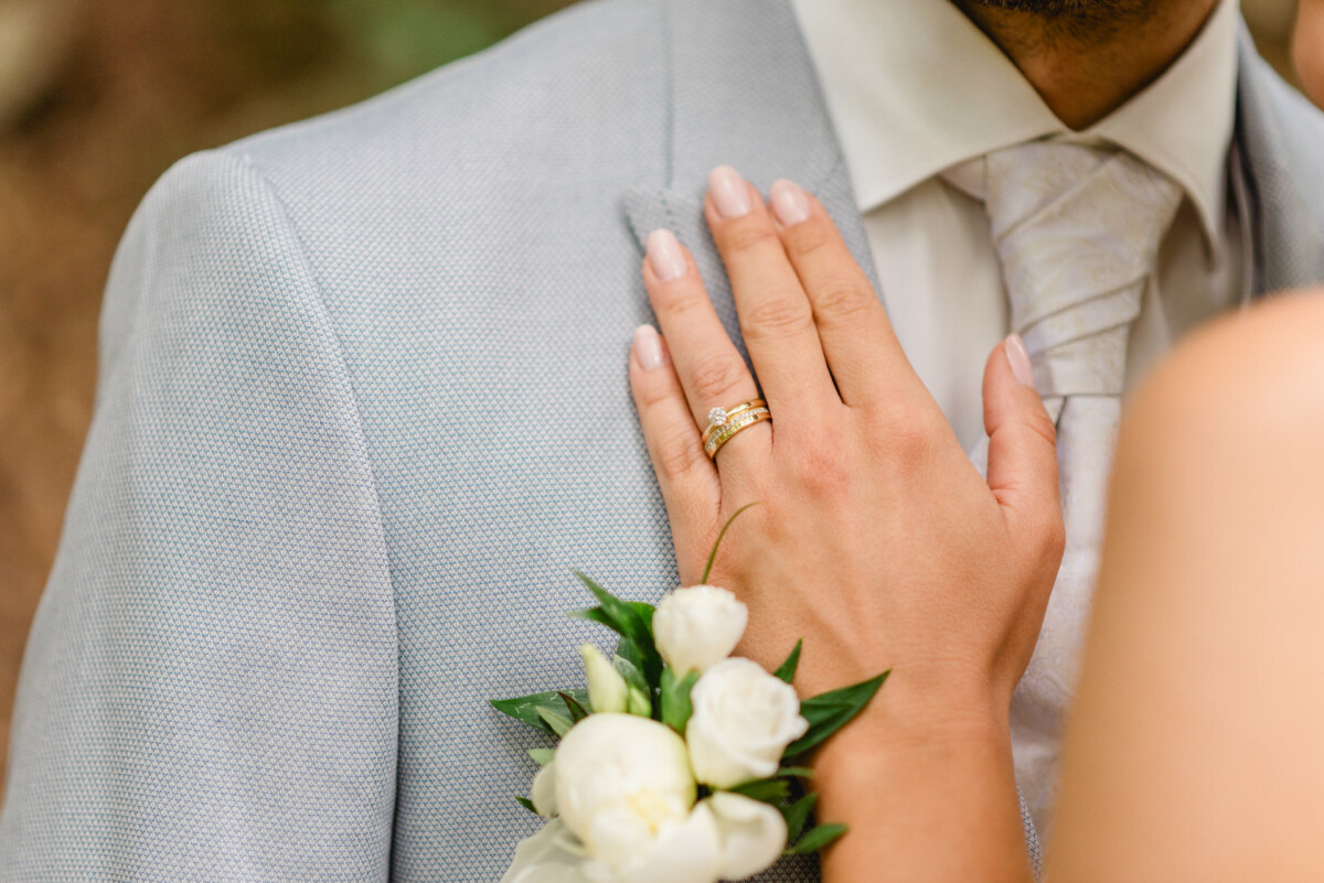 Die Hand einer Frau mit einem Ehering ruht auf der Brust eines Mannes im hellgrauen Anzug mit einer weißen Ansteckblume mit Blumenmotiv.