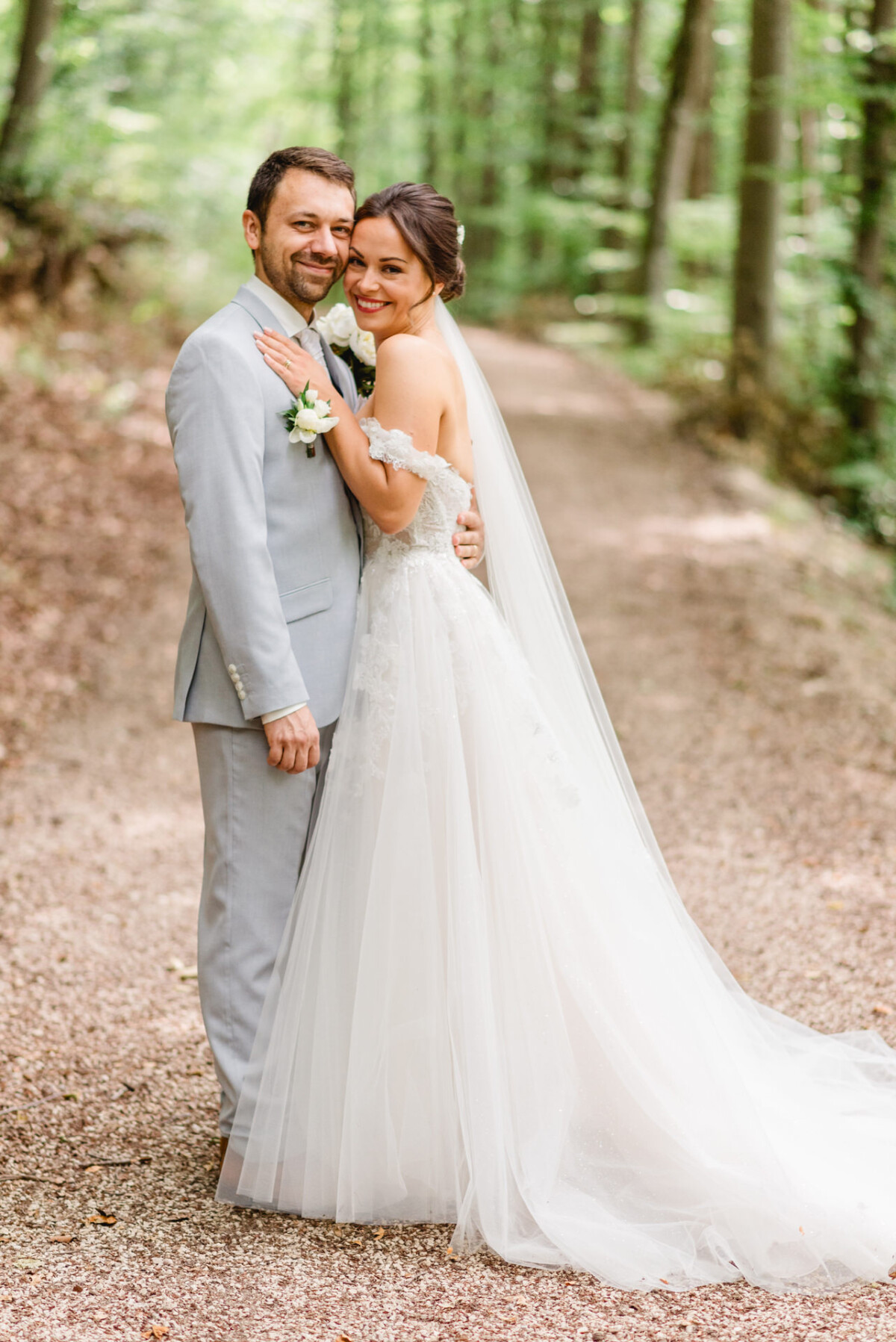 Ein Paar im Hochzeitskleid lächelt und umarmt sich auf einem Waldweg. Die Braut trägt ein weißes Kleid und einen Schleier, und der Bräutigam trägt einen hellgrauen Anzug. Im Hintergrund säumen Bäume den Weg.