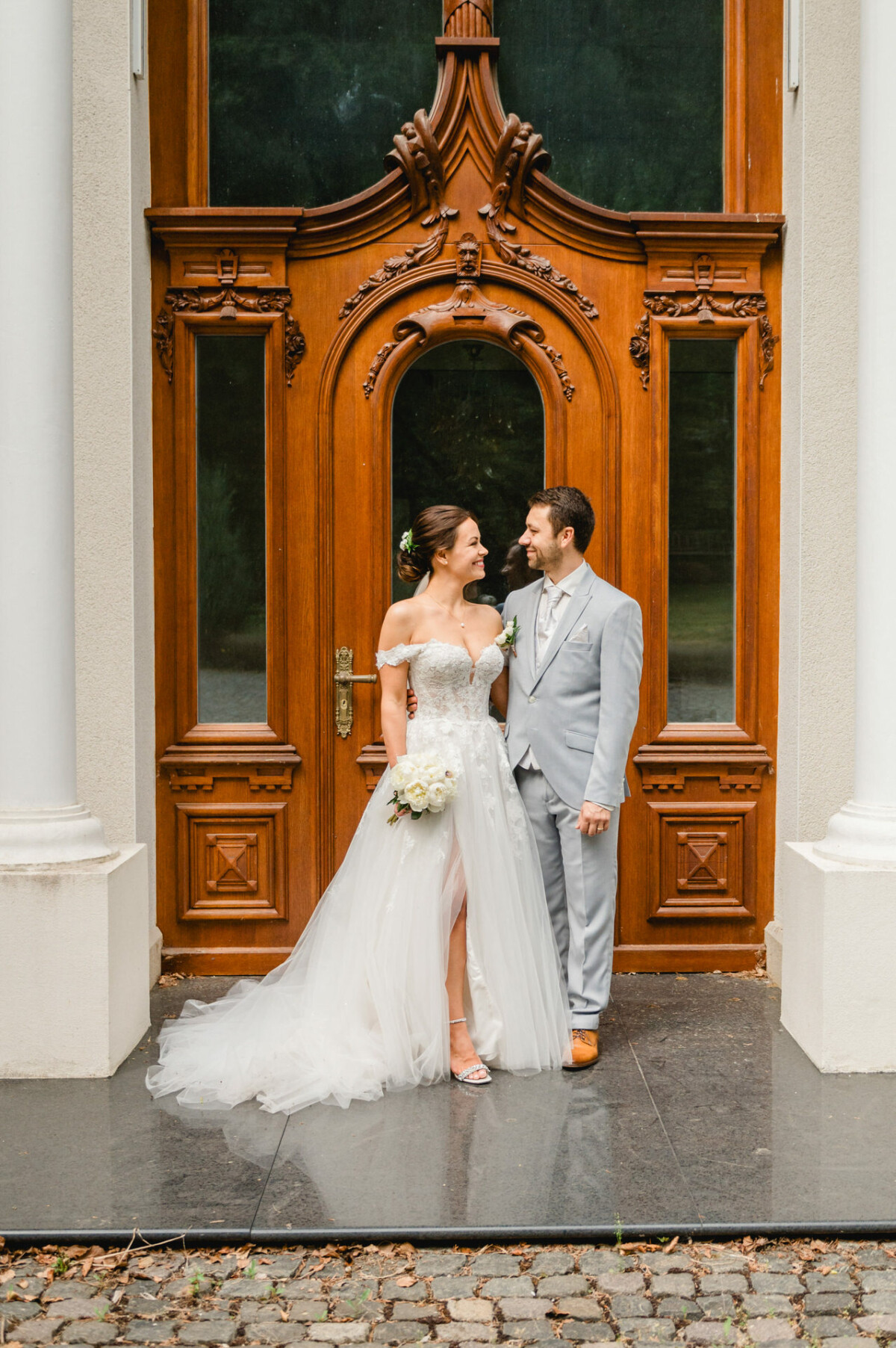 Braut und Bräutigam stehen vor einer verzierten Holztür. Die Braut trägt ein weißes Kleid und hält einen Blumenstrauß, der Bräutigam trägt einen hellgrauen Anzug.