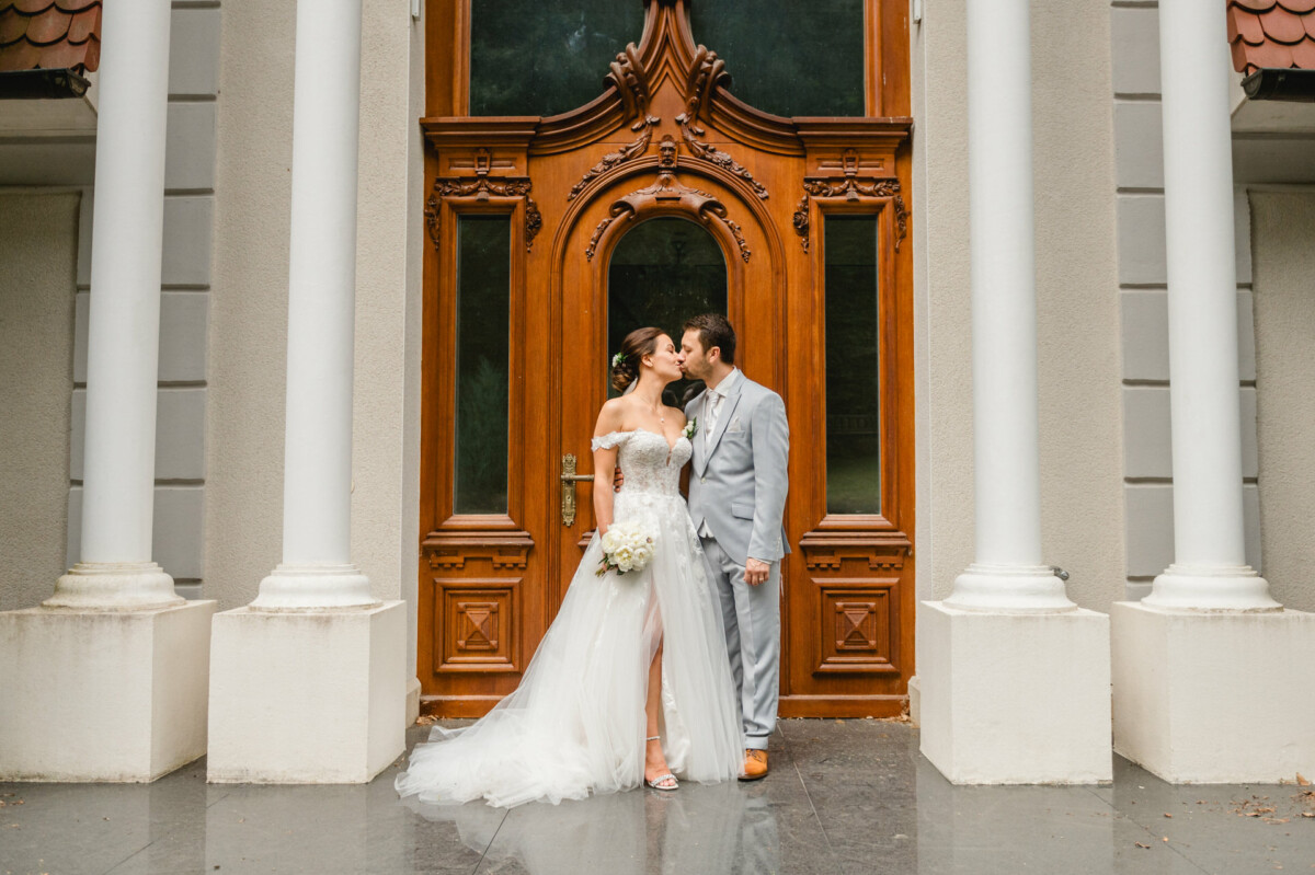 Eine Braut im weißen Kleid und ein Bräutigam im grauen Anzug küssen sich vor einer verzierten Holztür mit weißen Säulen.