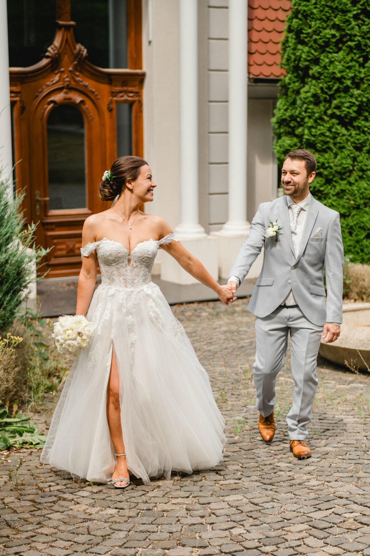 Eine Braut im weißen Kleid und ein Bräutigam im grauen Anzug halten Händchen und lächeln einander an, während sie auf einem Kopfsteinpflasterweg spazieren.