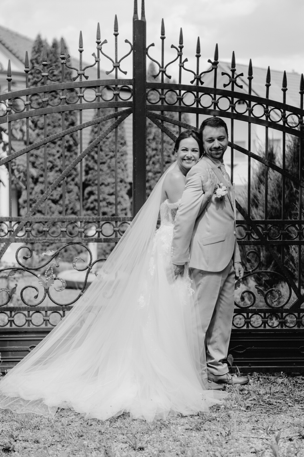 Braut und Bräutigam stehen lächelnd Rücken an Rücken vor einem verzierten schmiedeeisernen Tor, der Schleier der Braut fällt herab.