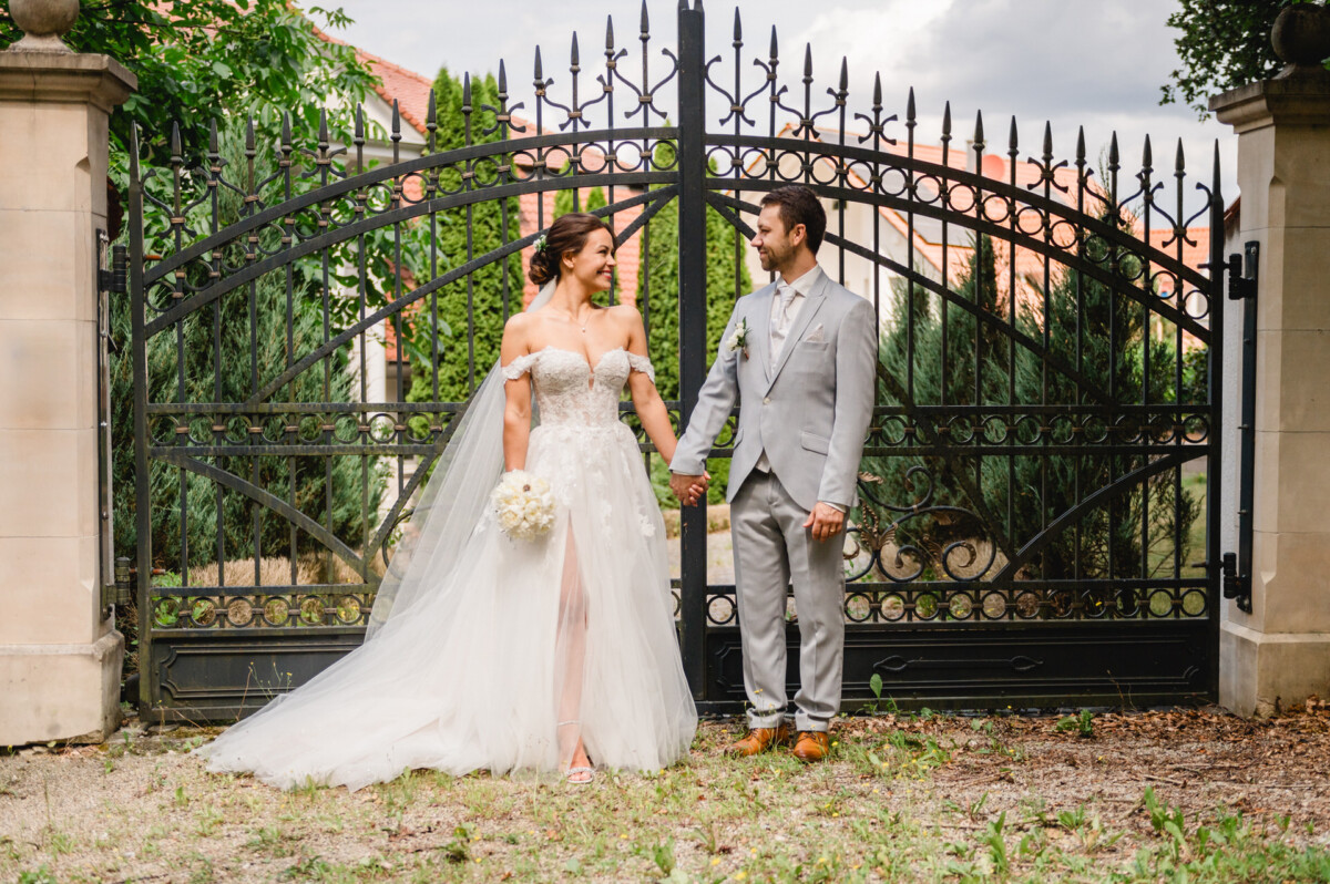 Eine Braut im weißen Kleid und ein Bräutigam im grauen Anzug stehen Händchen haltend vor einem kunstvoll verzierten schwarzen Tor.