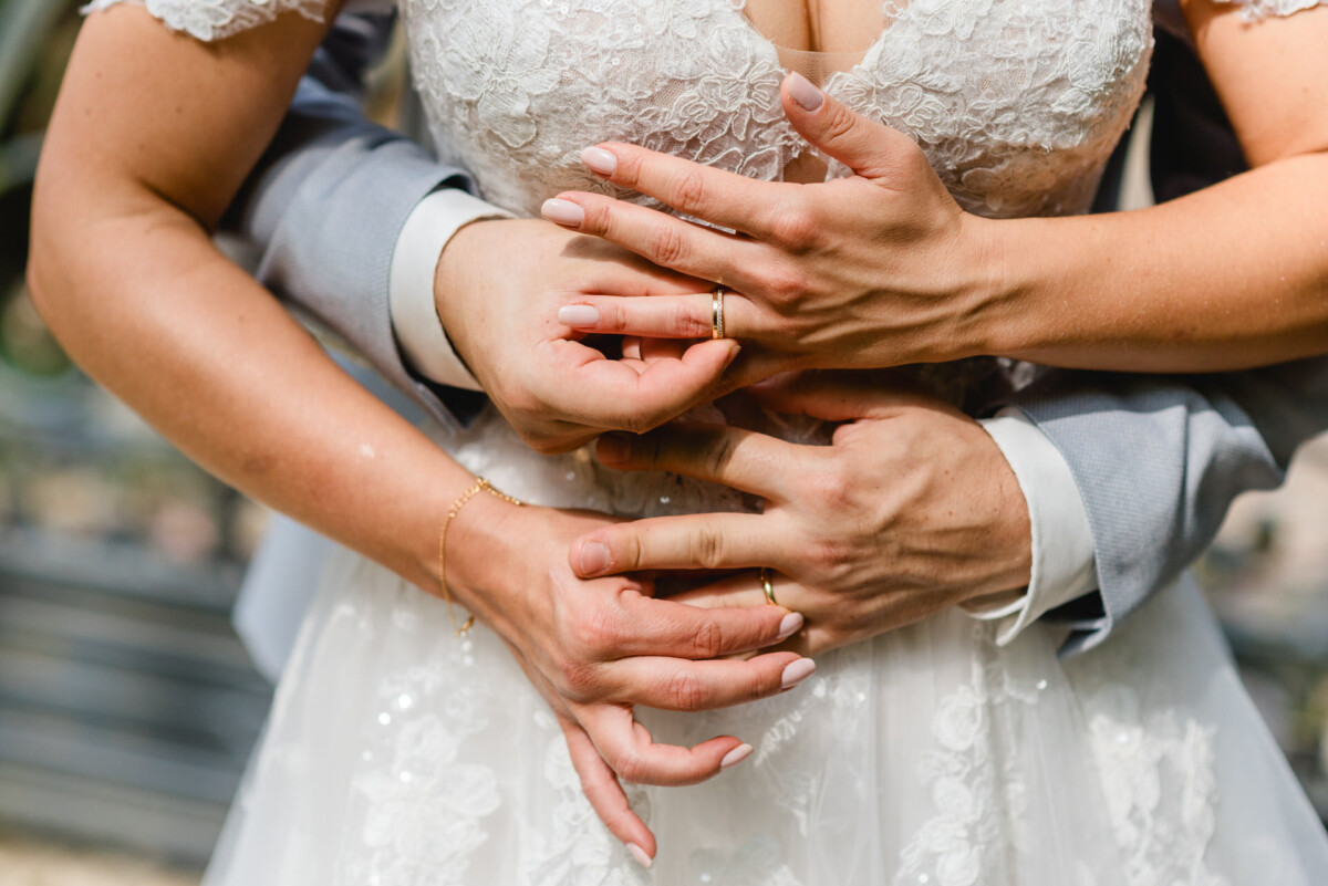 Ein Paar im Hochzeitskleid umarmt sich und zeigt seine Eheringe an den ineinander verschlungenen Händen.