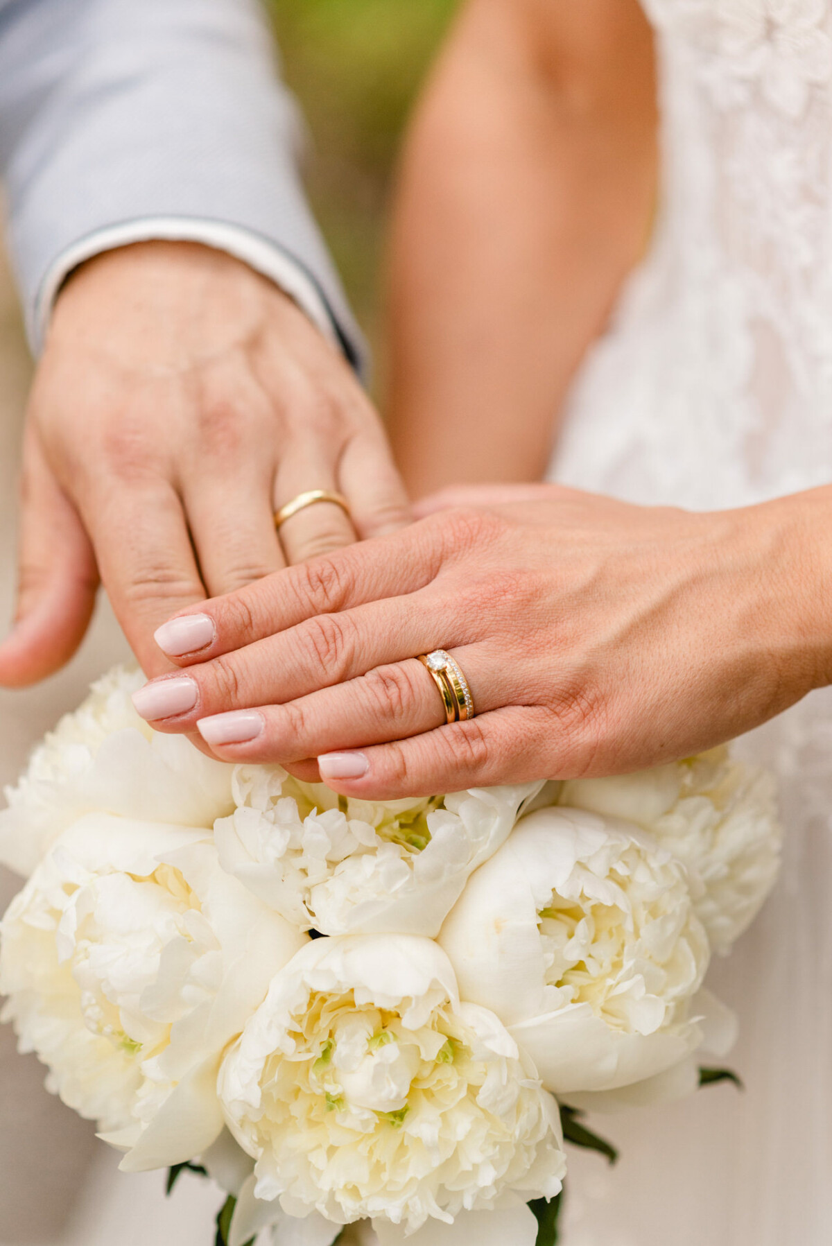 Nahaufnahme der Hände eines Paares; beide tragen Eheringe und ruhen auf einem Strauß weißer Blumen.