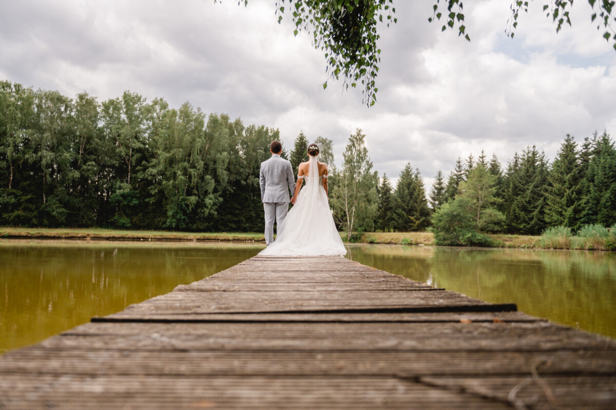 Braut und Bräutigam stehen auf einem Holzsteg mit Blick auf einen Teich, umgeben von Bäumen und einem bewölkten Himmel.
