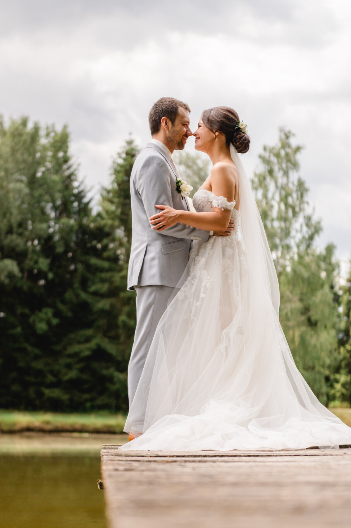 Braut und Bräutigam lächeln sich auf einem Holzsteg an, umgeben von Bäumen, während die Braut ein wallendes Kleid und einen Schleier trägt.