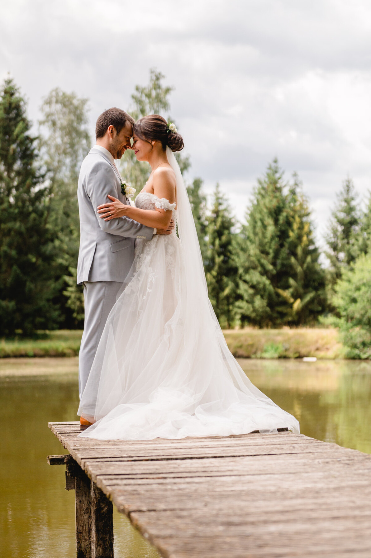 Braut und Bräutigam umarmen sich auf einem Holzsteg über einem ruhigen See, umgeben von üppigen Bäumen, unter einem bewölkten Himmel.