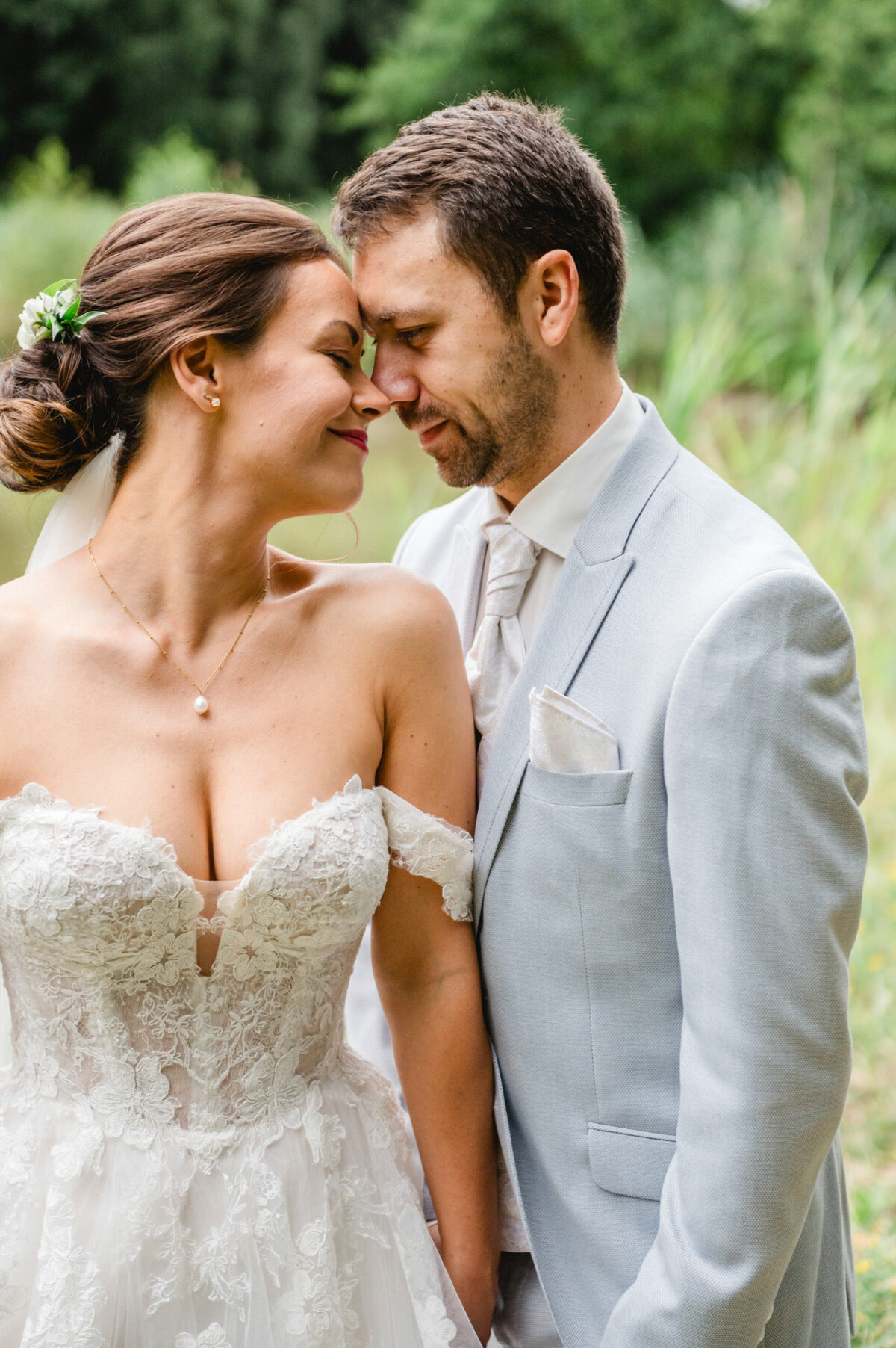 Braut und Bräutigam stehen eng beieinander im Freien, die Braut trägt ein weißes Spitzenkleid und der Bräutigam einen hellgrauen Anzug. Sie lächeln und berühren ihre Stirnen, umgeben von Grün.