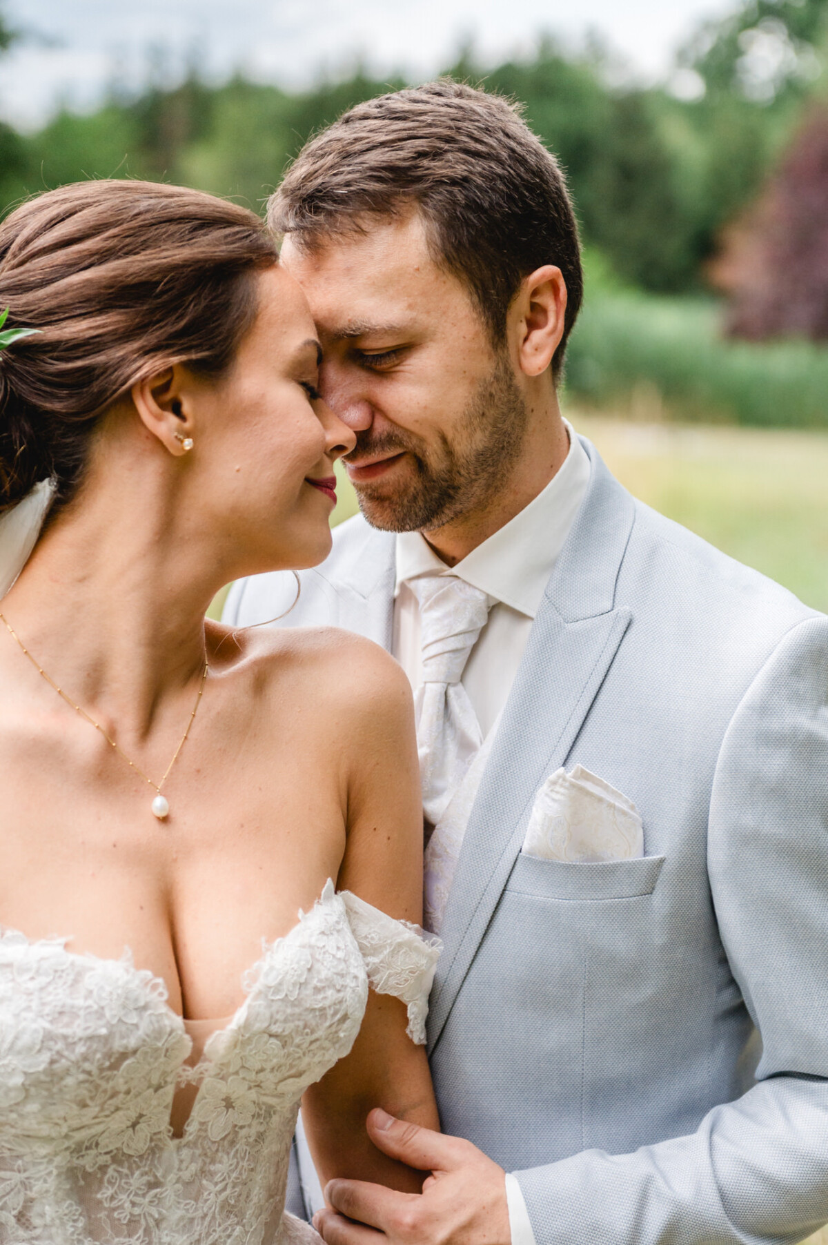 Braut und Bräutigam umarmen sich im Freien, beide lächeln und haben die Augen geschlossen. Die Braut trägt ein Spitzenkleid und der Bräutigam einen hellen Anzug. Im Hintergrund ist Grün zu sehen.