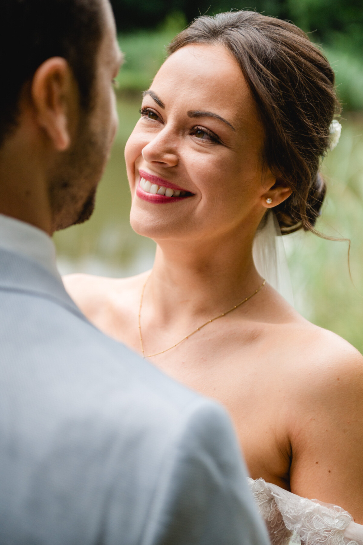 Eine Braut lächelt ihren Bräutigam im Freien in einer natürlichen Umgebung an. Sie trägt ein trägerloses Kleid und einen Schleier.