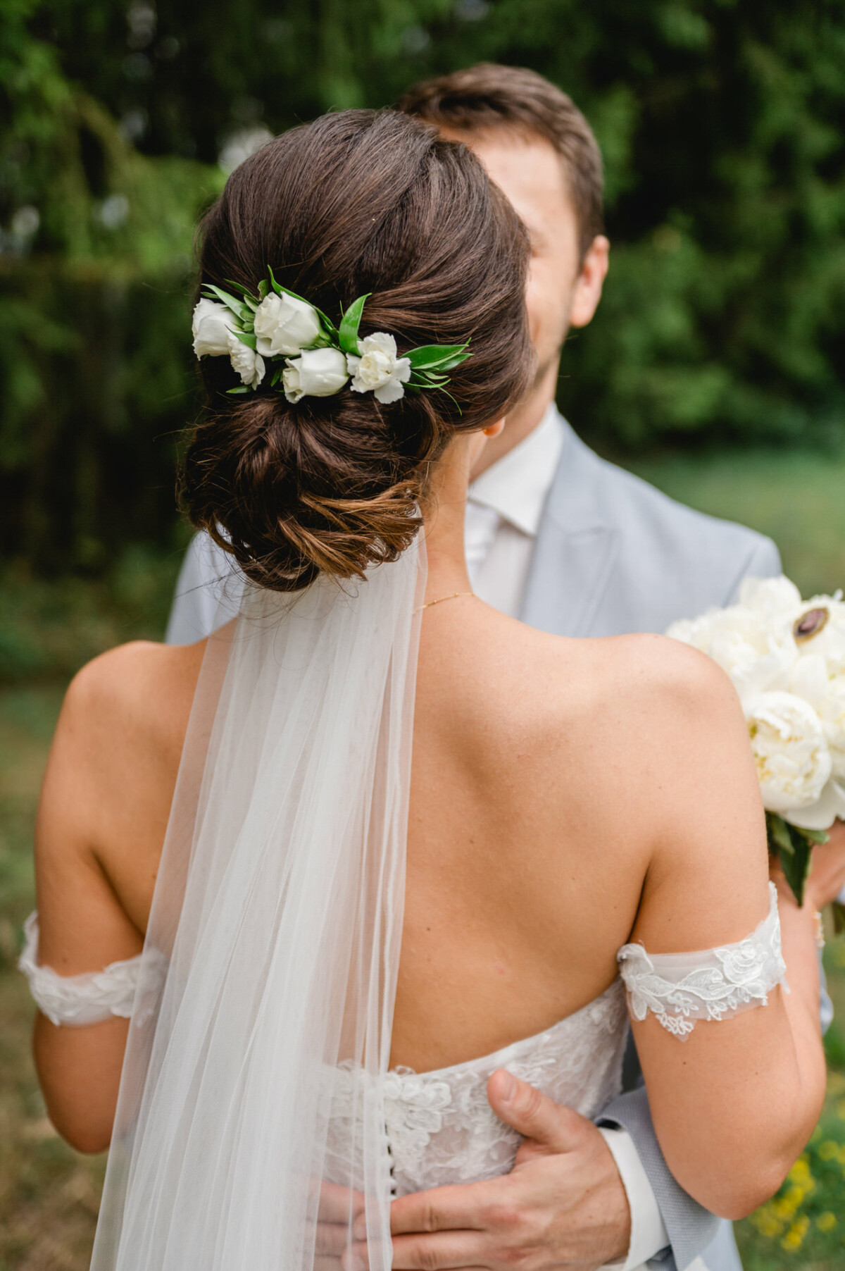 Braut mit Blumenhaarschmuck und Schleier wird von einem Bräutigam im Anzug umarmt. Sie hält einen Strauß weißer Blumen. Außenkulisse mit viel Grün im Hintergrund.