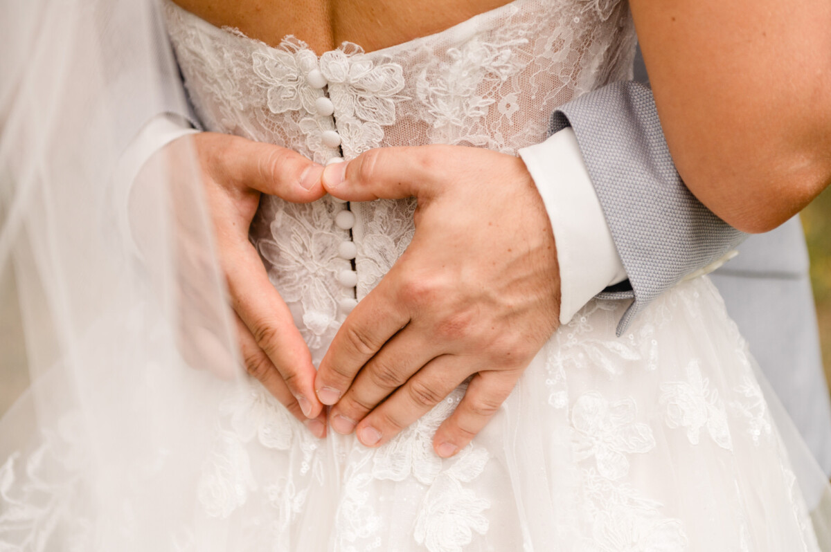 Nahaufnahme eines Bräutigams, der mit seinen Händen auf der Rückseite des Spitzenhochzeitskleides einer Braut ein Herz formt.