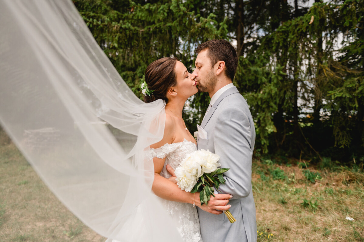 Braut und Bräutigam küssen sich im Freien. Die Braut hält einen Strauß weißer Blumen in der Hand und ihr Schleier weht hinter ihr her.