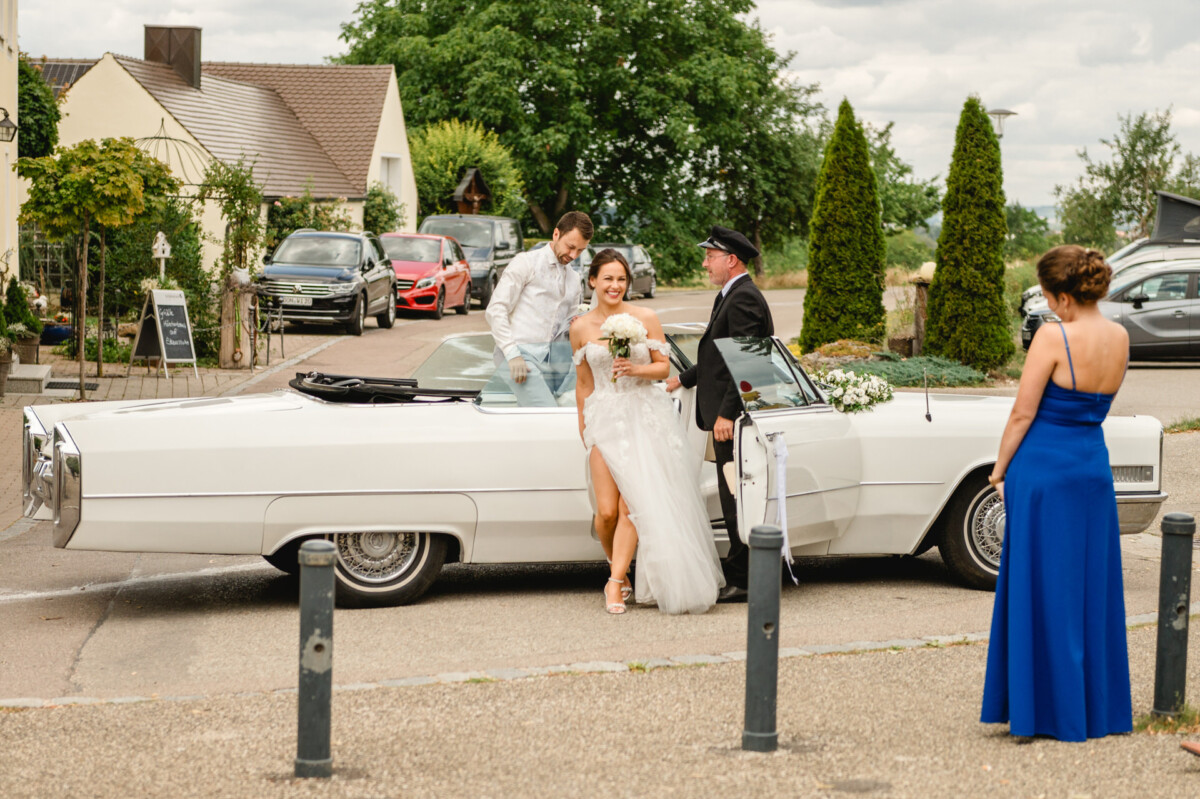 Braut und Bräutigam steigen aus einem weißen Cabrio, während eine Frau in einem blauen Kleid zusieht. Eine weitere Person steht in der Nähe.