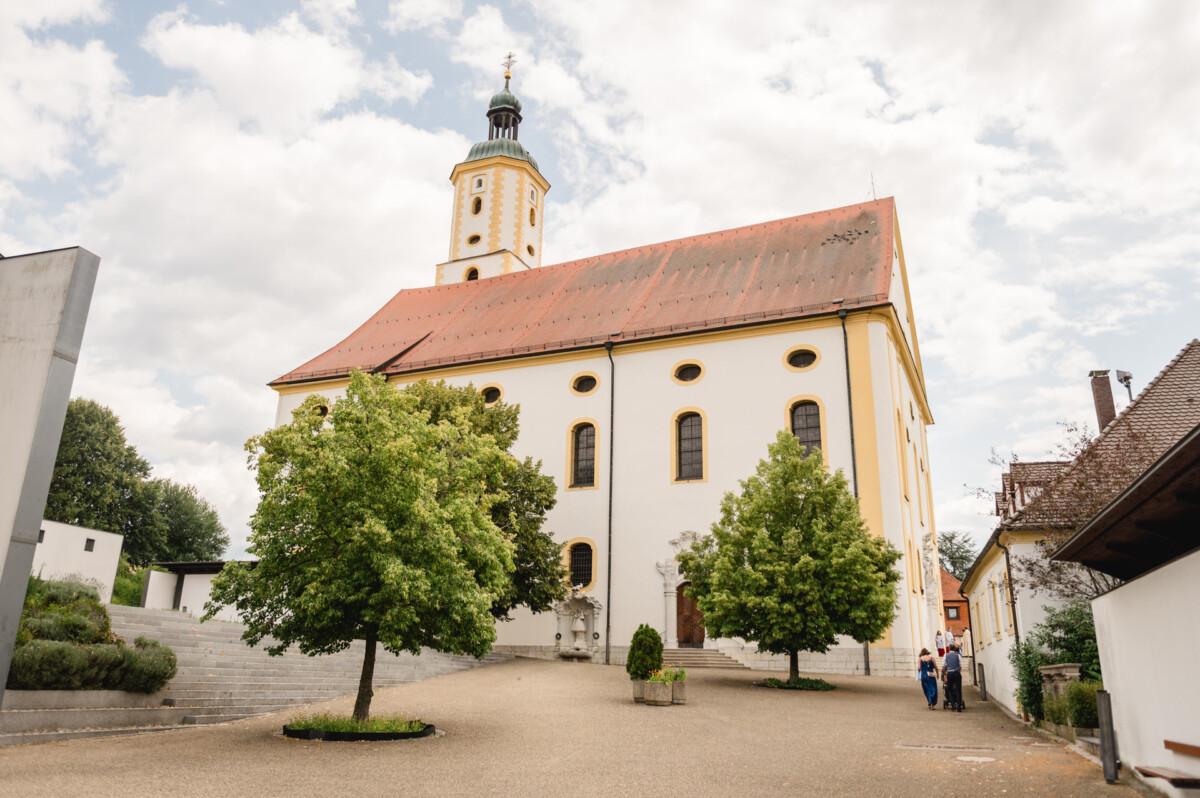 Eine weiße Kirche mit rotem Dach und Turm steht zwischen Bäumen und einem Steinpfad unter einem bewölkten Himmel.