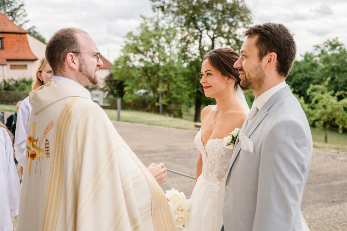 Ein Paar im Hochzeitskleid steht im Freien einem Geistlichen gegenüber. Die Braut hält einen Blumenstrauß und der Bräutigam trägt einen hellgrauen Anzug. Im Hintergrund sind Bäume und Gebäude zu sehen.
