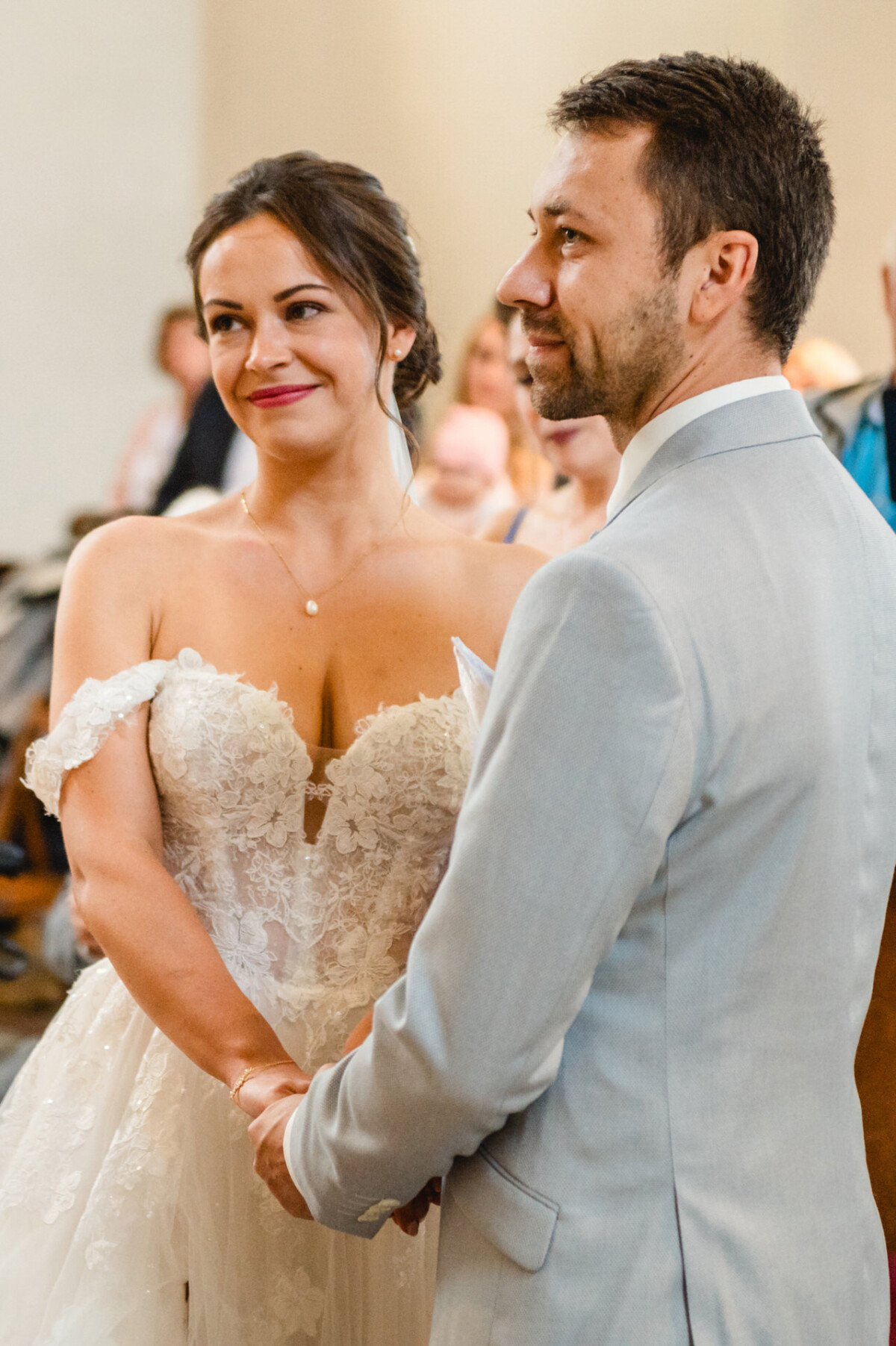 Braut und Bräutigam stehen während einer Hochzeitszeremonie Händchen haltend da. Die Braut trägt ein Spitzenkleid und der Bräutigam einen hellen Anzug.