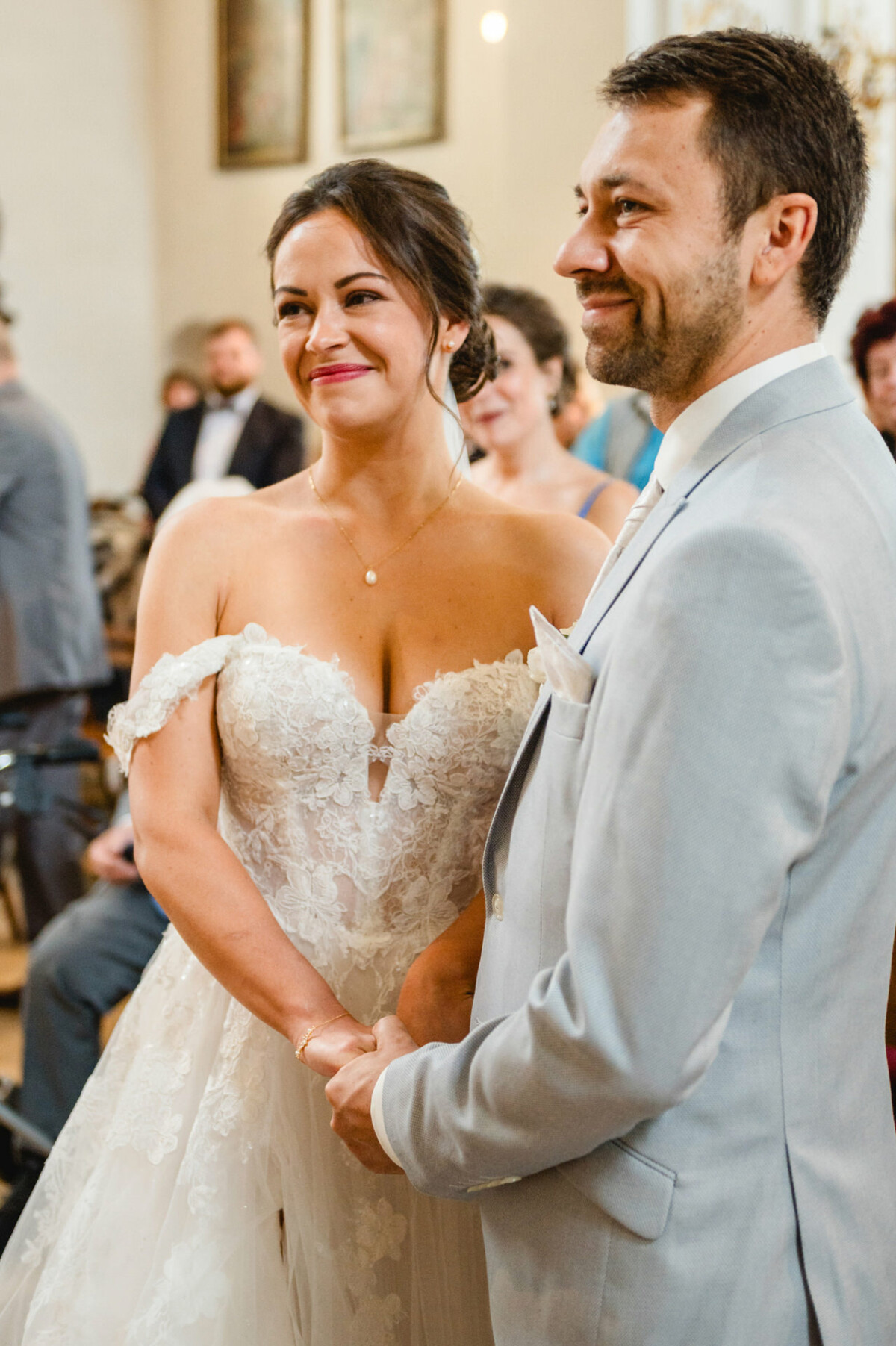 Braut und Bräutigam stehen während ihrer Hochzeitszeremonie Händchen haltend da und lächeln beide. Die Braut trägt ein weißes Kleid und der Bräutigam einen hellgrauen Anzug.