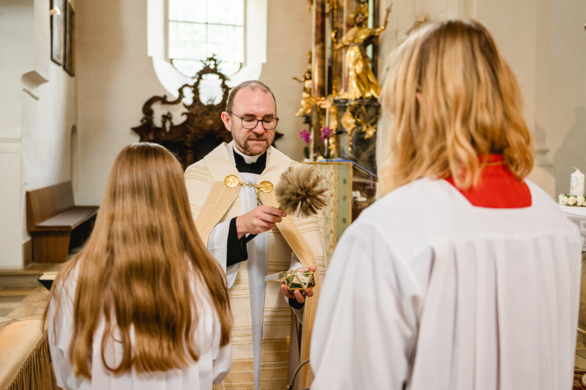 Ein Priester im liturgischen Gewand führt in einer Kirche vor zwei Menschen in weißen Gewändern ein Segensritual mit einem Pinsel durch.