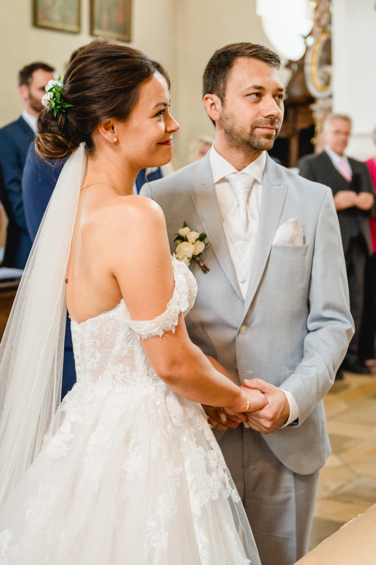 Braut und Bräutigam halten während einer Hochzeitszeremonie Händchen. Die Braut trägt ein weißes Kleid mit langem Schleier und der Bräutigam einen hellgrauen Anzug mit weißer Krawatte.