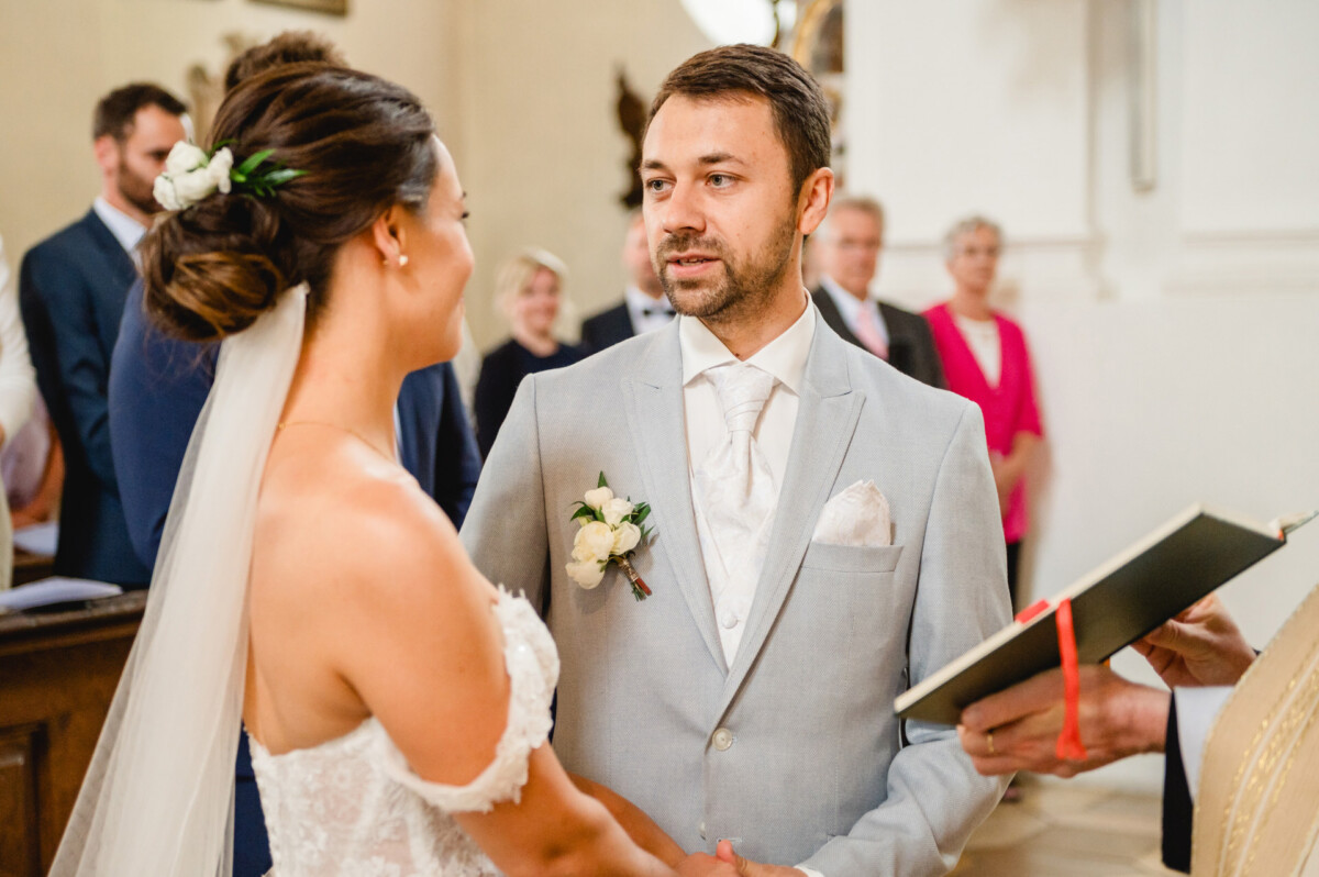 Braut und Bräutigam stehen bei einer Hochzeitszeremonie zusammen und halten sich an den Händen. Der Bräutigam trägt einen hellgrauen Anzug und die Braut ein weißes Kleid mit Schleier, umgeben von Gästen.