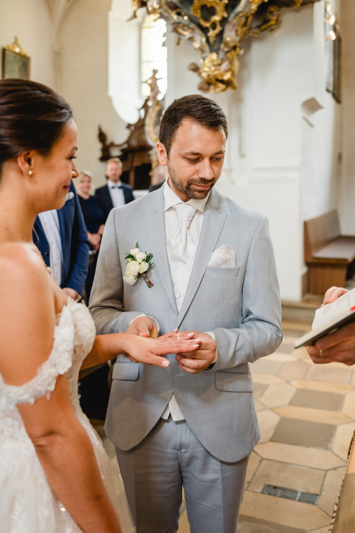 Braut und Bräutigam tauschen bei einer kirchlichen Zeremonie Ringe aus. Der Bräutigam trägt einen hellgrauen Anzug und die Braut hält den Ring. In der Nähe hält eine Person ein offenes Buch.